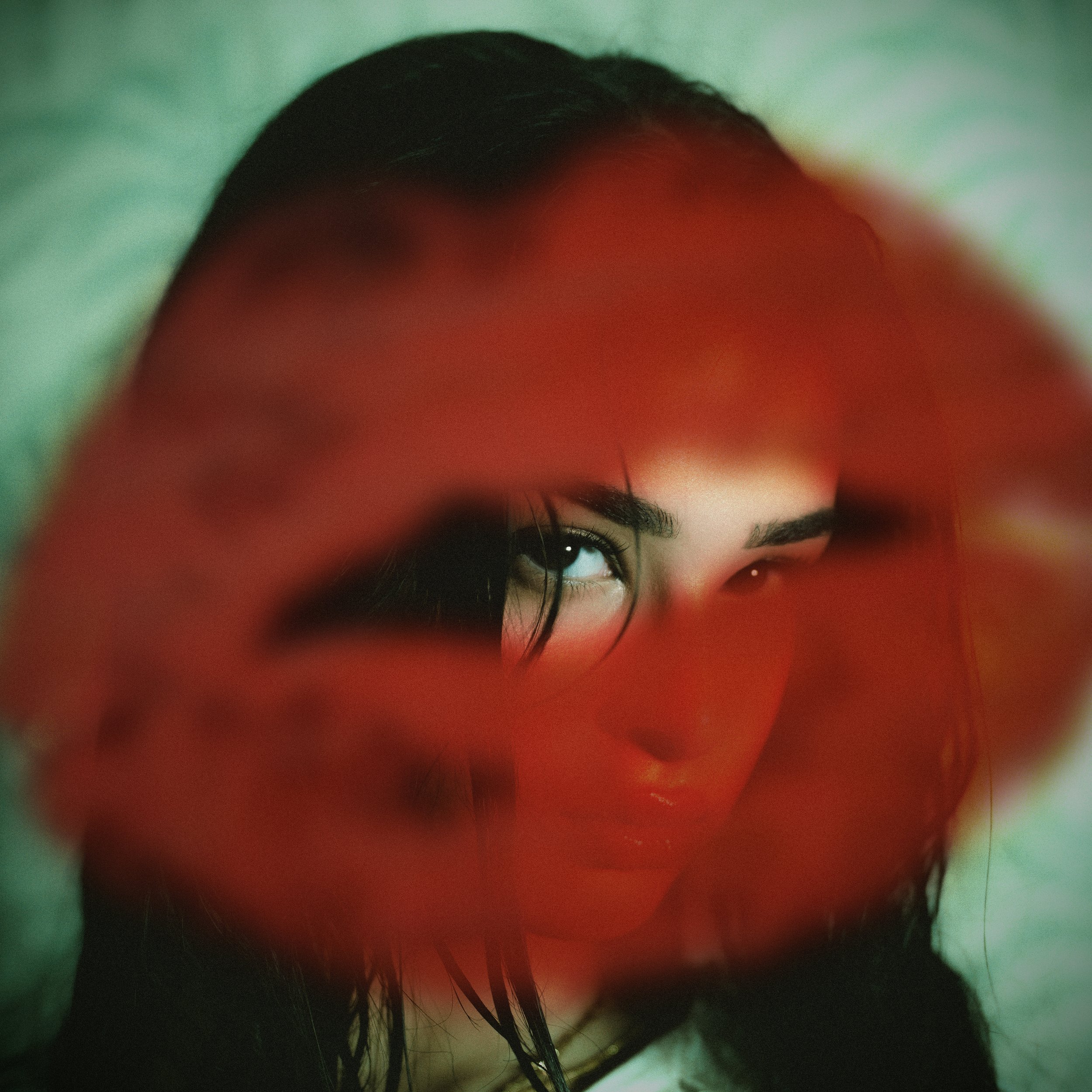 A woman with dark hair and eyebrows peeks through lipstick marks, with only part of her face visible, showing her eyes and forehead.