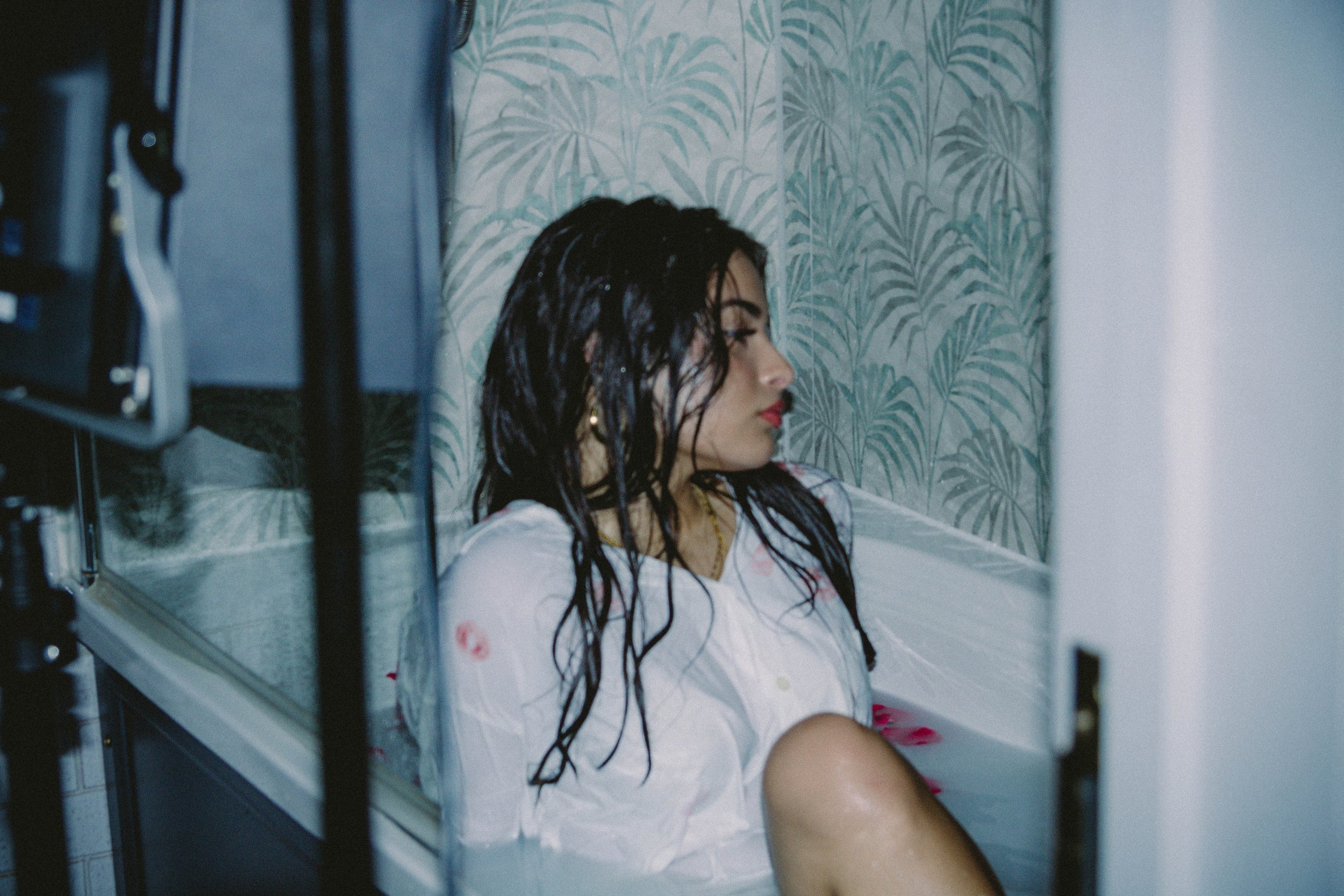 A woman with wet dark hair and red lipstick is sitting in a bathtub filled with water, looking down with her eyes closed, in a bathroom with tropical wallpaper.