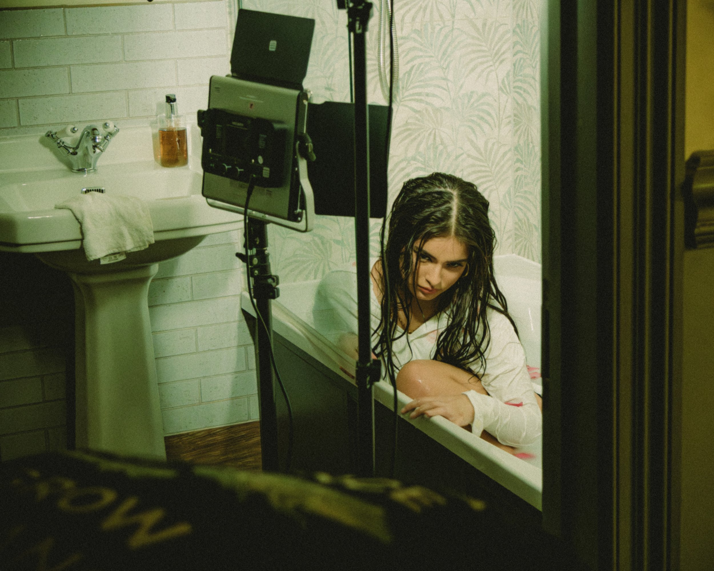 A woman with wet hair and a white shirt sitting in a bathtub with water, looking at the camera with an intense expression. The scene is being filmed with a camera setup visible in the foreground, inside a room with white brick walls, a sink with a bottle of liquid soap, and patterned wallpaper.
