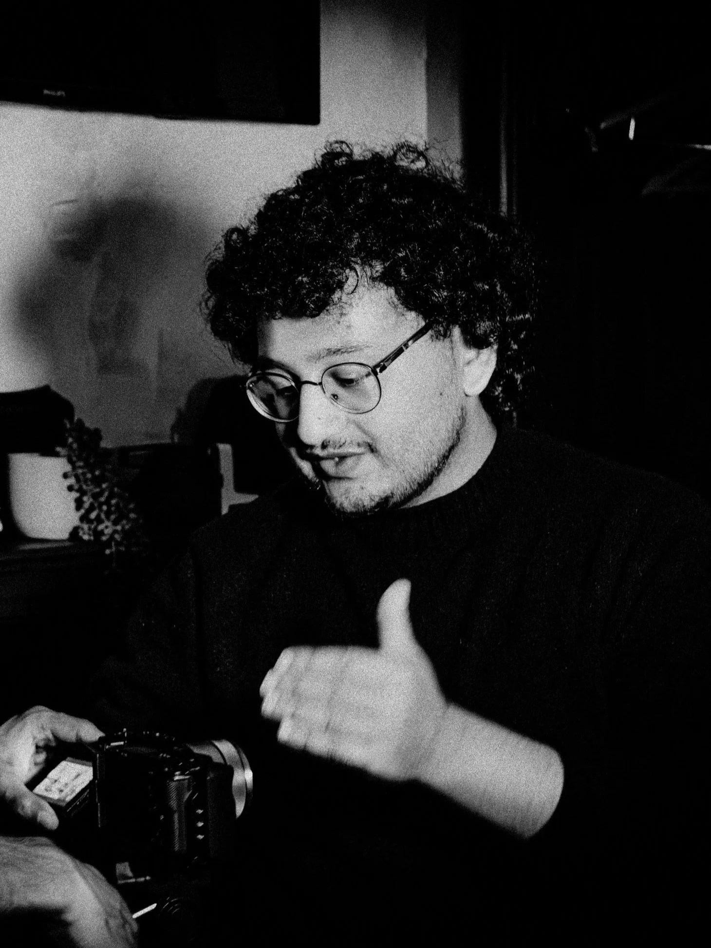 A young man with curly hair and glasses, wearing a dark sweater, looking at a camera in his hands.