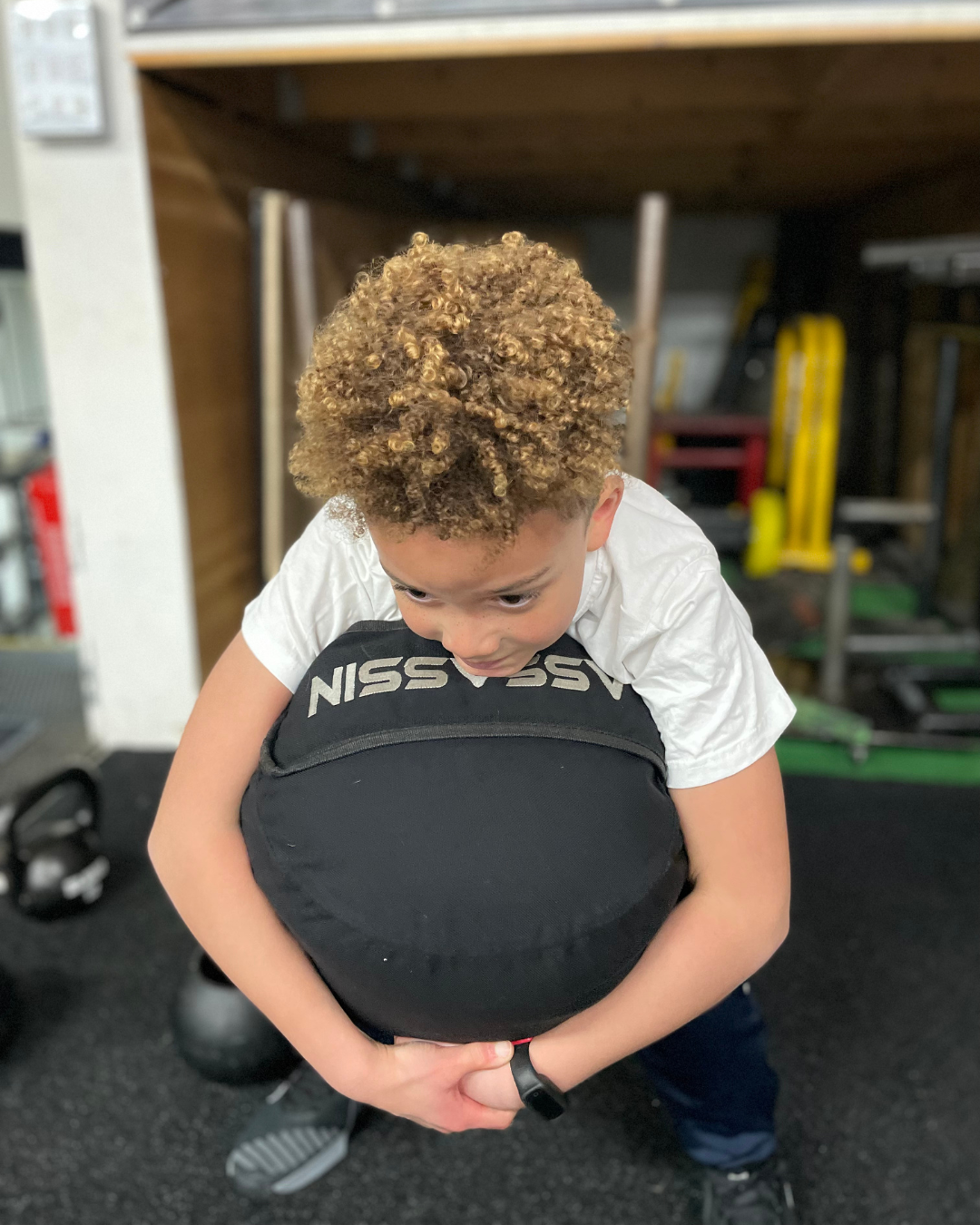 Two young boys participating in a strongman event, lifting large black medicine balls in an outdoor sports area, with tents and banners in the background.