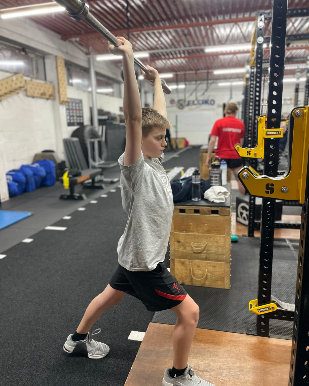 A young boy in a gym squatting with a wooden barbell on his shoulders, surrounded by fitness equipment.