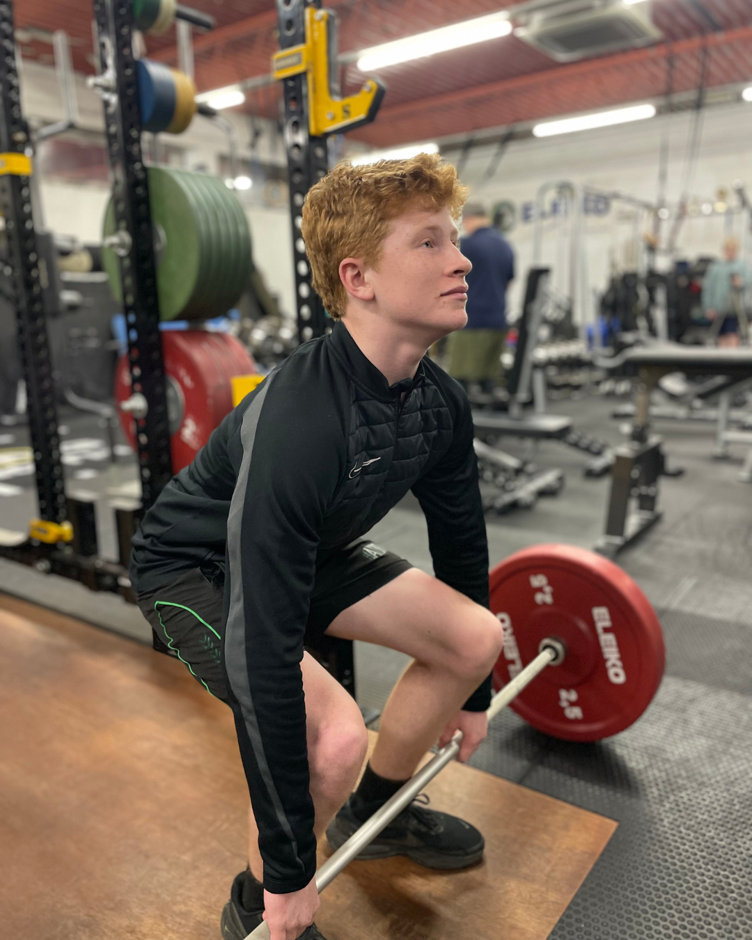 Young boy lifting a barbell in a gym, wearing a Liverpool football jersey.
