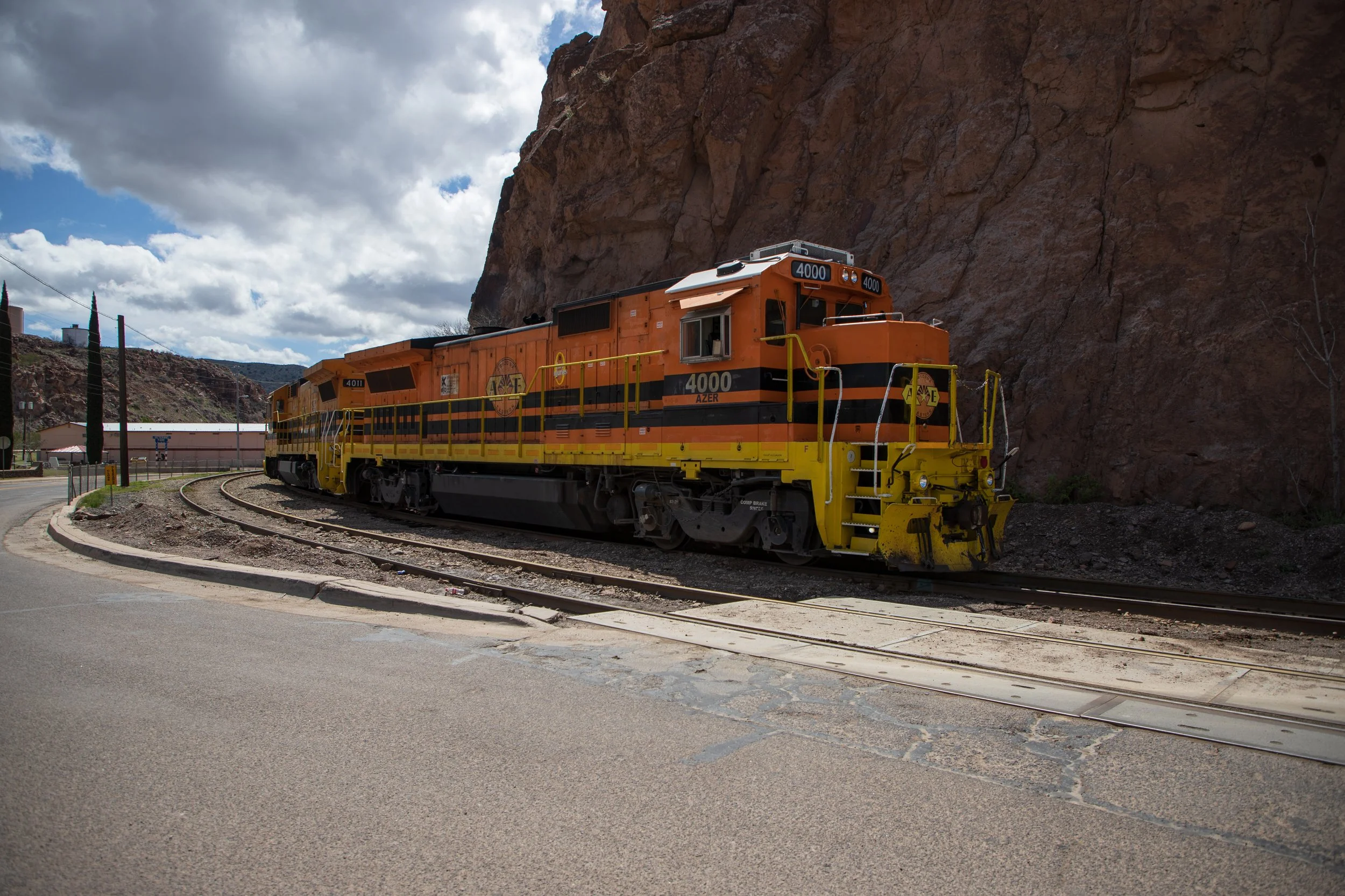 Orange and yellow locomotive train on tracks beside rocky cliff under cloudy sky.