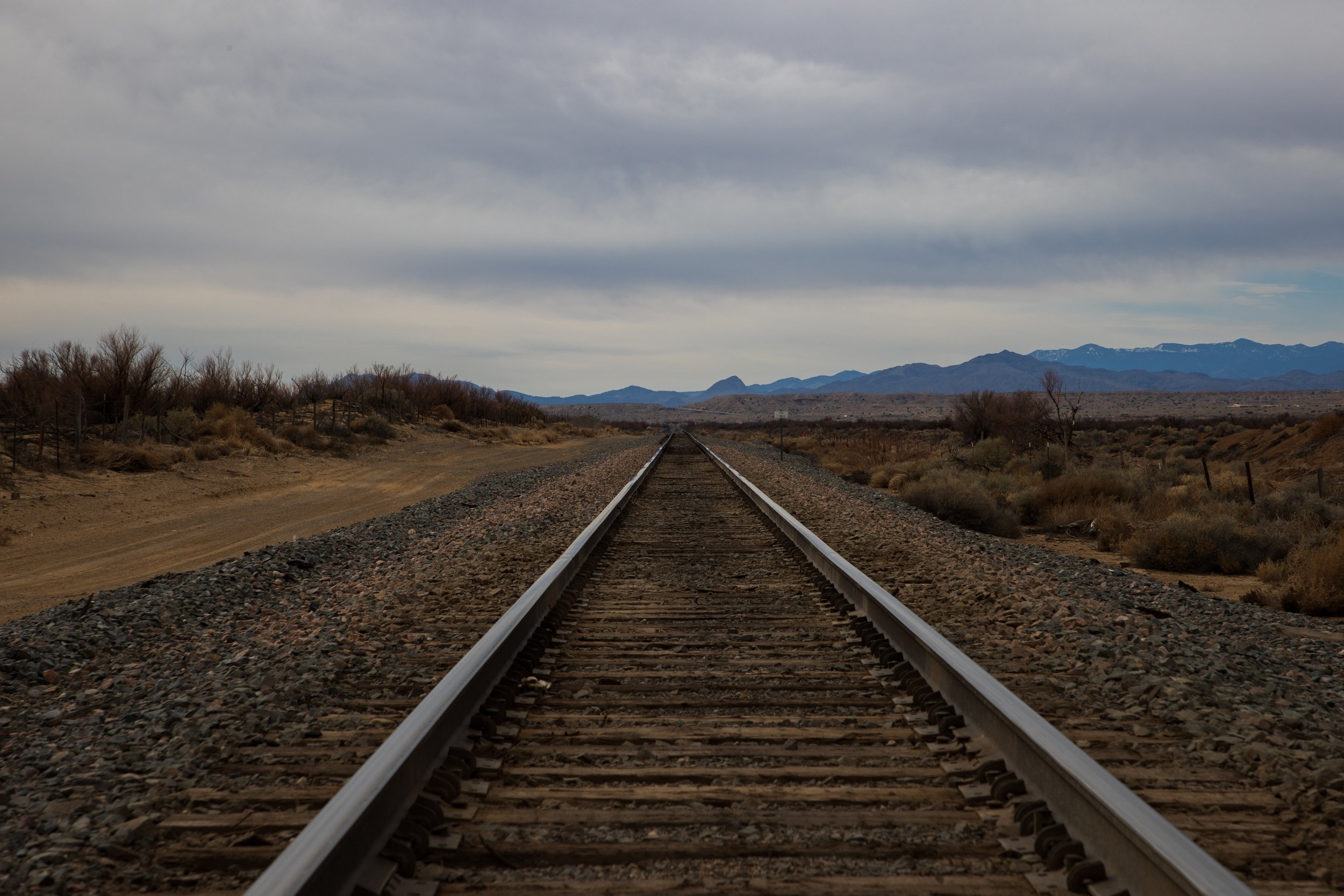 Straight railway tracks in a desert landscape with distant mountains under a cloudy sky.