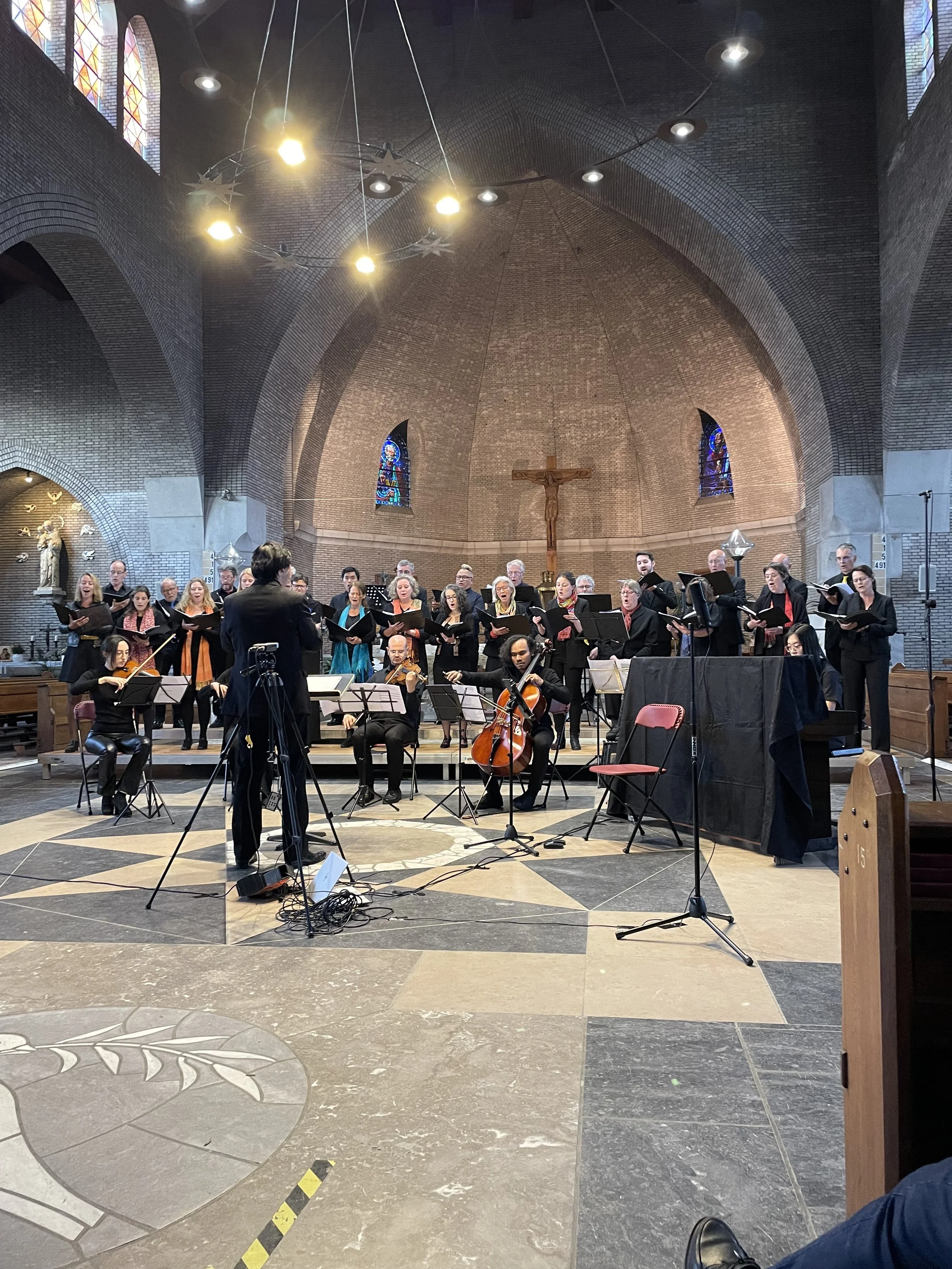 A choir and orchestra performing in a church interior with a crucifix and stained glass windows.