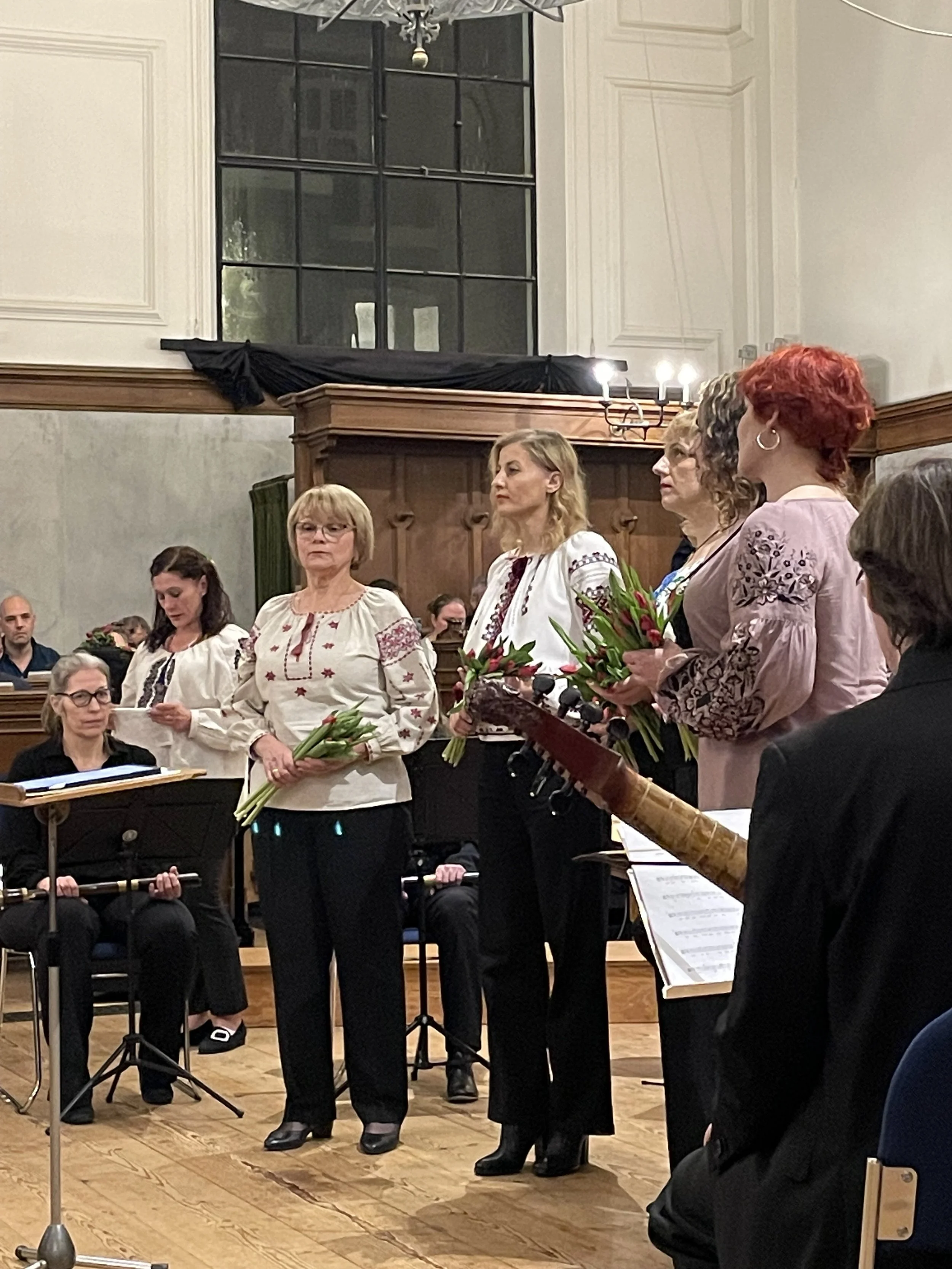 A group of women standing indoors, holding flowers and dressed in traditional embroidered clothing. They appear to be part of a choir or musical ensemble. Seated musicians with sheet music are visible in the background. The setting is a room with wooden flooring and large windows.