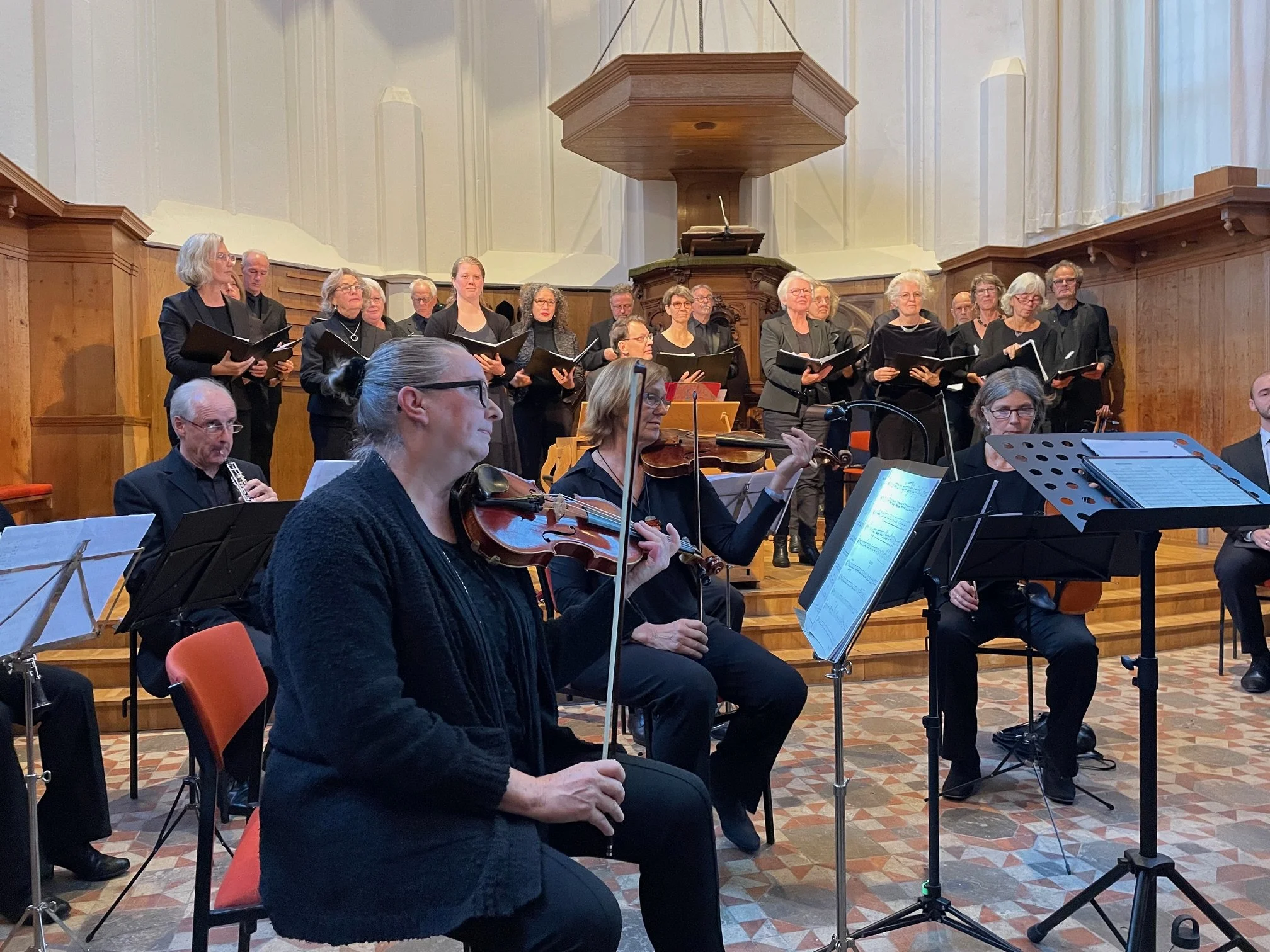 An orchestra and choir performing in a church setting, with musicians playing violins and wind instruments in the foreground, and choir members holding music sheets in the background.