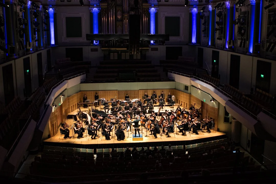 A large orchestra on stage at the National Concert Hall in Dublin. There are blue lights around the walls, and the members of the orchestra are all wearing black,.