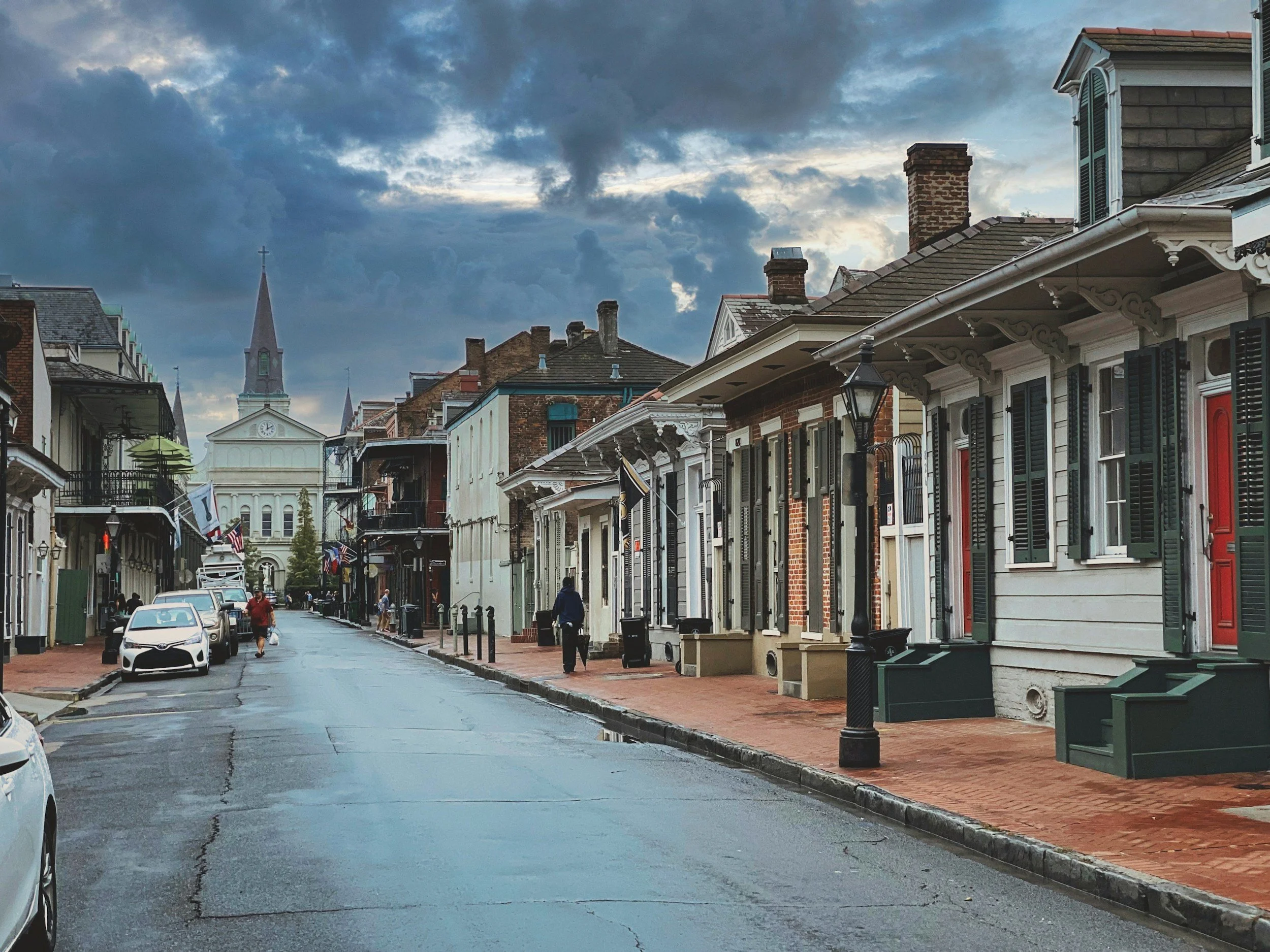 A street with historic buildings and a church steeple in the background, under a cloudy sky.