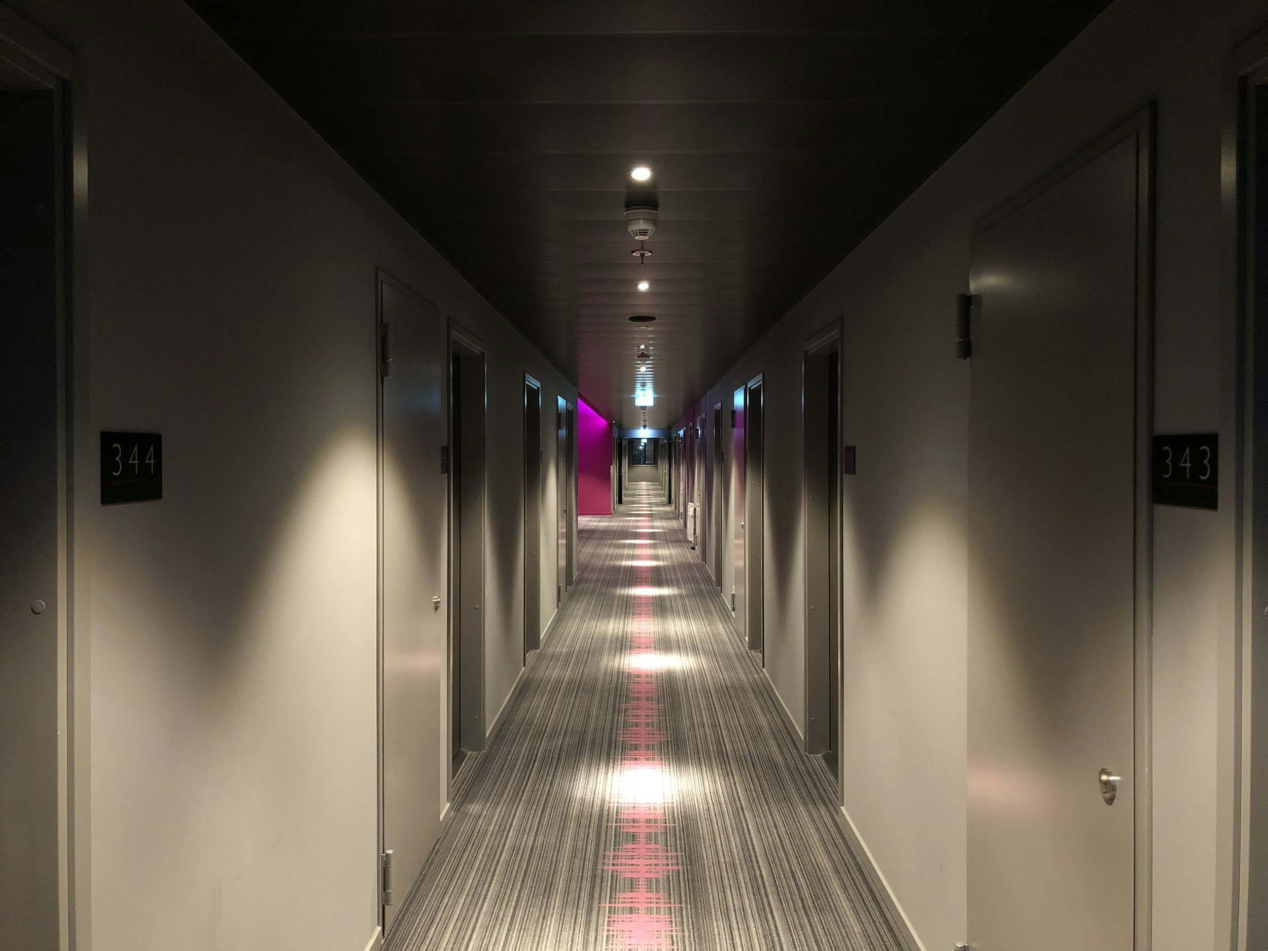 Empty hotel corridor with closed doors, numbered 344 and 343, illuminated by recessed ceiling lights, with colorful accent lighting at the end of the hallway.