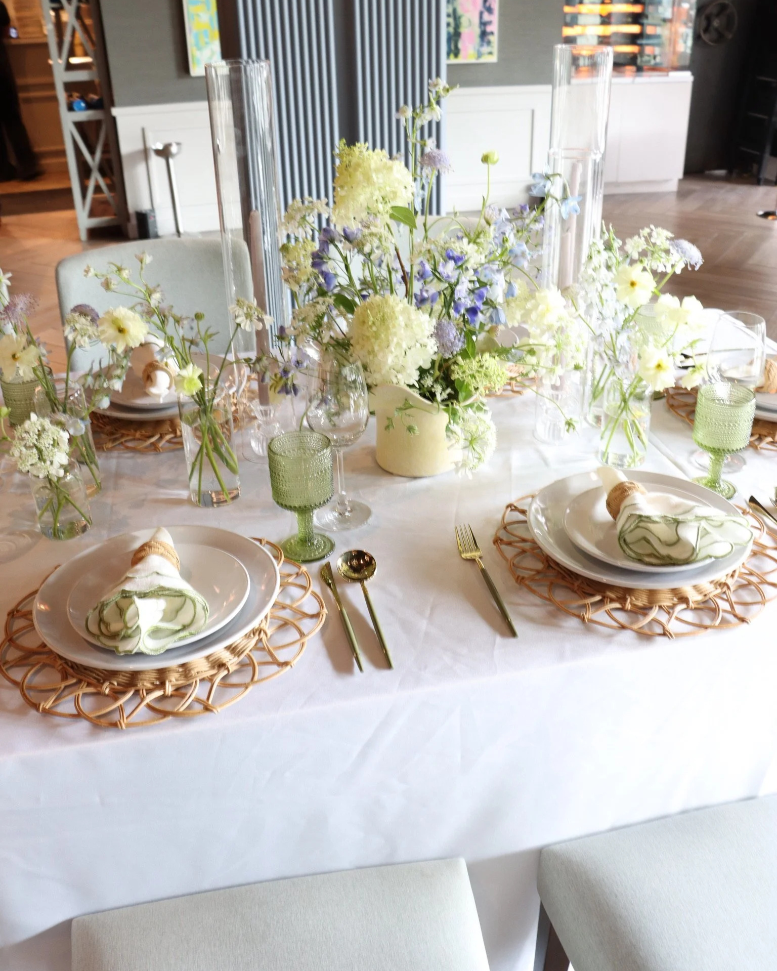 Elegant table setting with white plates, green napkins, golden utensils, and a large floral centerpiece of white and purple flowers in a cream vase, surrounded by glassware and greenery.