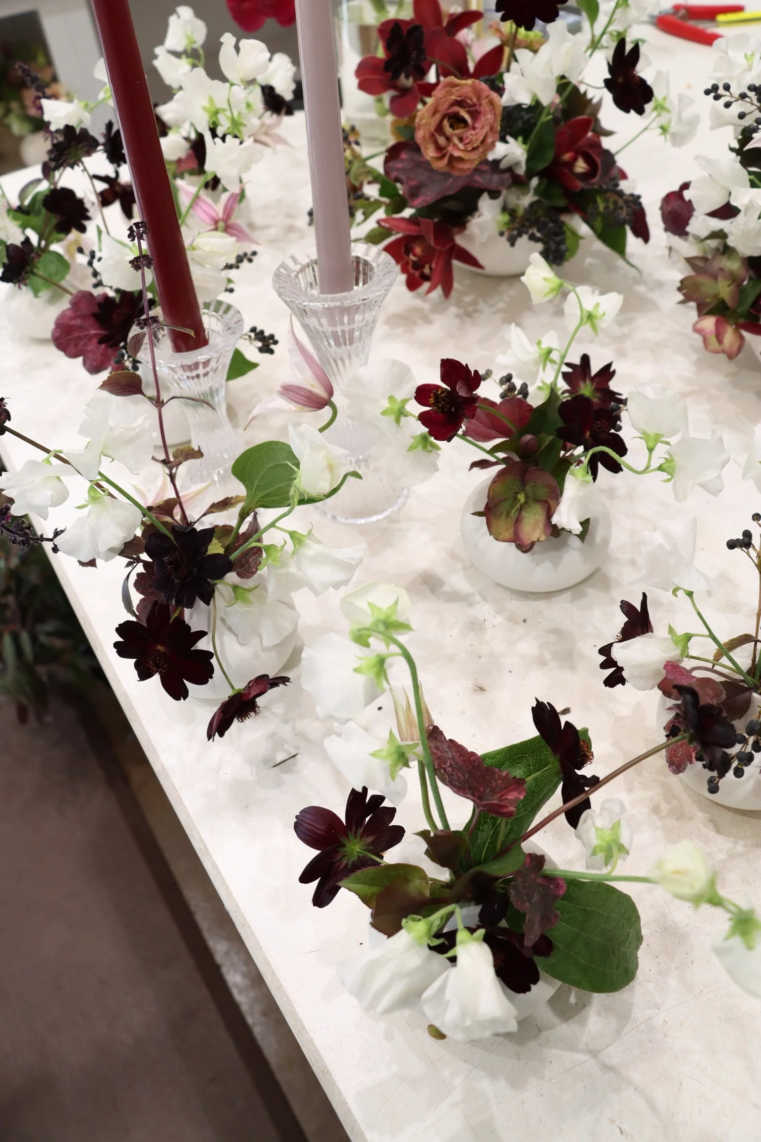 Table decorated with vases and flowers, including white, dark red, and some pinkish blossoms, along with two tall candle holders with candles.
