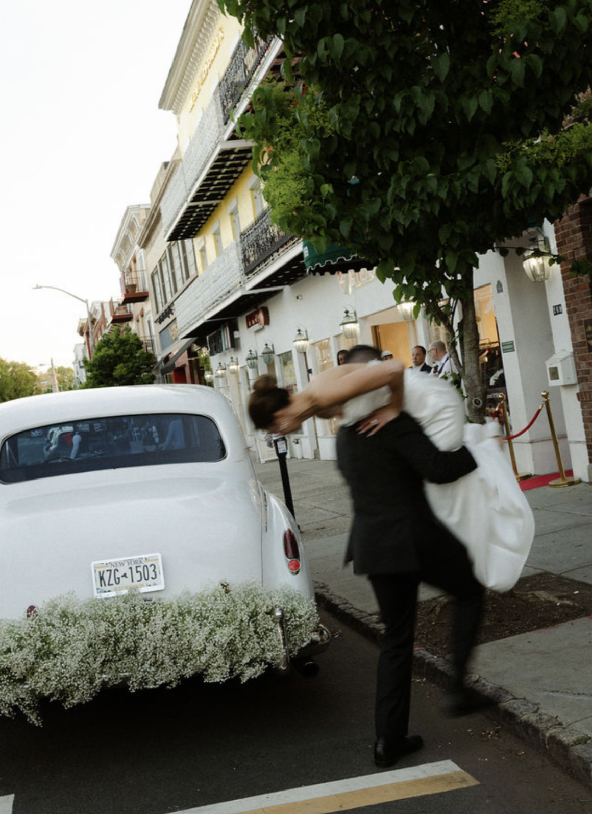 A person in a black suit is lifting and carrying a bride in a white wedding dress on a city sidewalk. There is a white vintage car decorated with flowers parked nearby, and a multi-story building with balconies in the background. Trees and sidewalk elements are also visible.