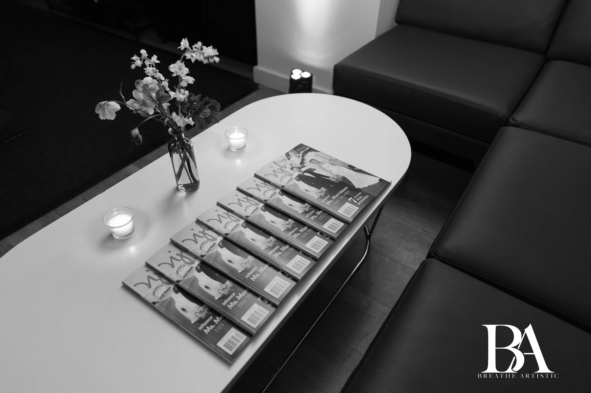 Table with multiple magazines, a vase with flowers, lit candles, and a small decorative object, in a living room setting.