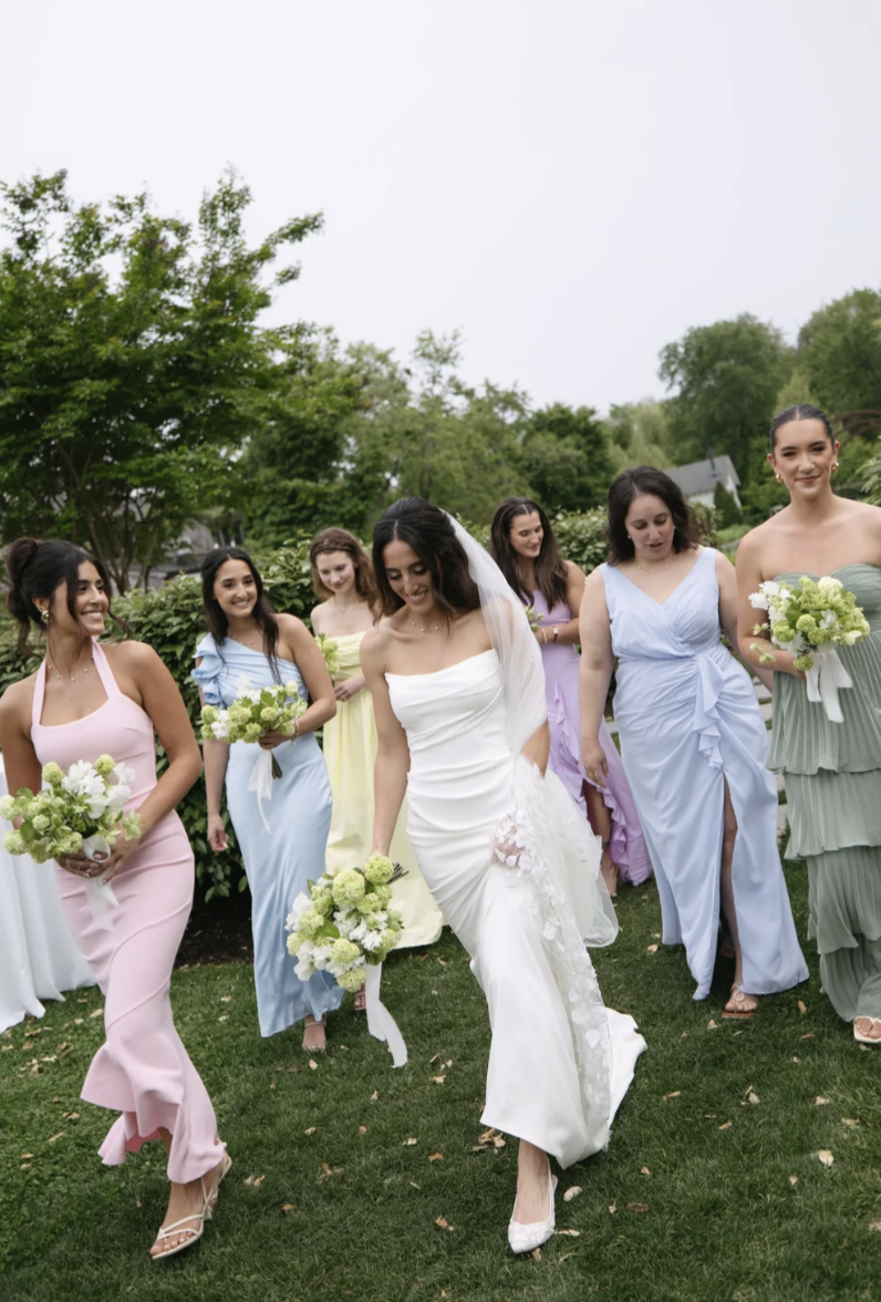 Bride and bridesmaids walking on grass outdoors, holding bouquets, in pastel dresses.