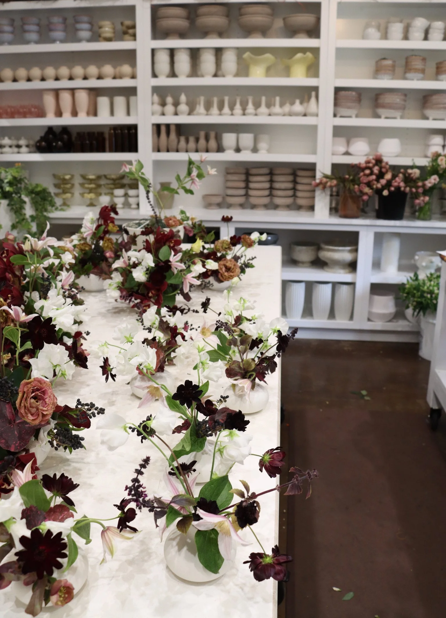 A workspace with a marble table featuring several floral arrangements in small white vases, with shelves of pottery and vases in the background.