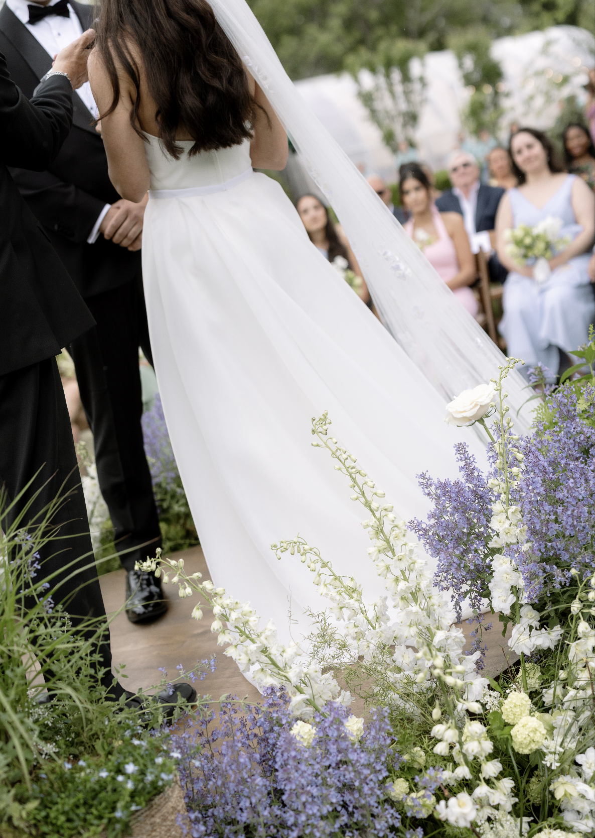 A bride in a white wedding dress and veil standing at the altar during a wedding ceremony, surrounded by purple and white flowers, with guests seated in the background.