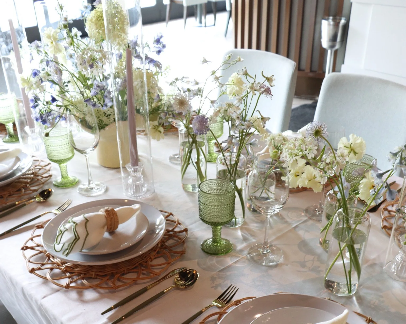 A dining table set with white plates, gold utensils, white napkins, and woven placemats. The table is decorated with various vases of white, purple, and green flowers, along with green glass goblets and candles.