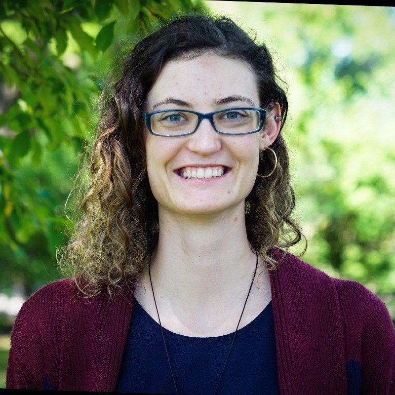 A young woman with wavy brown hair, wearing glasses, a dark blue top, a burgundy cardigan, and gold hoop earrings, standing outdoors with green trees in the background.