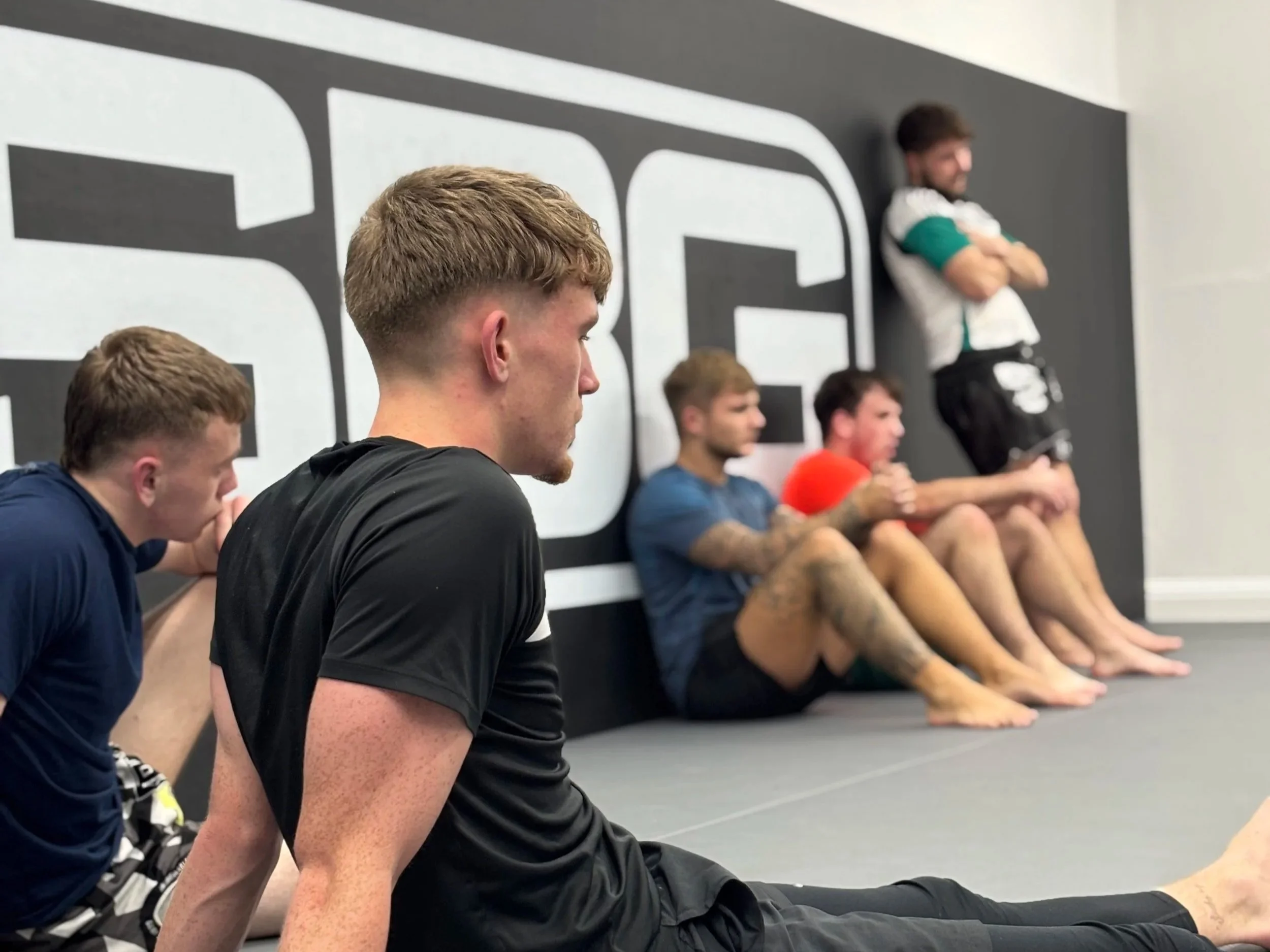 A group of five young men sitting on the floor in a gym, leaning against a wall with the letters 'SBG' in the background. One man in the foreground is stretching, while the others sit with crossed arms or relaxed.