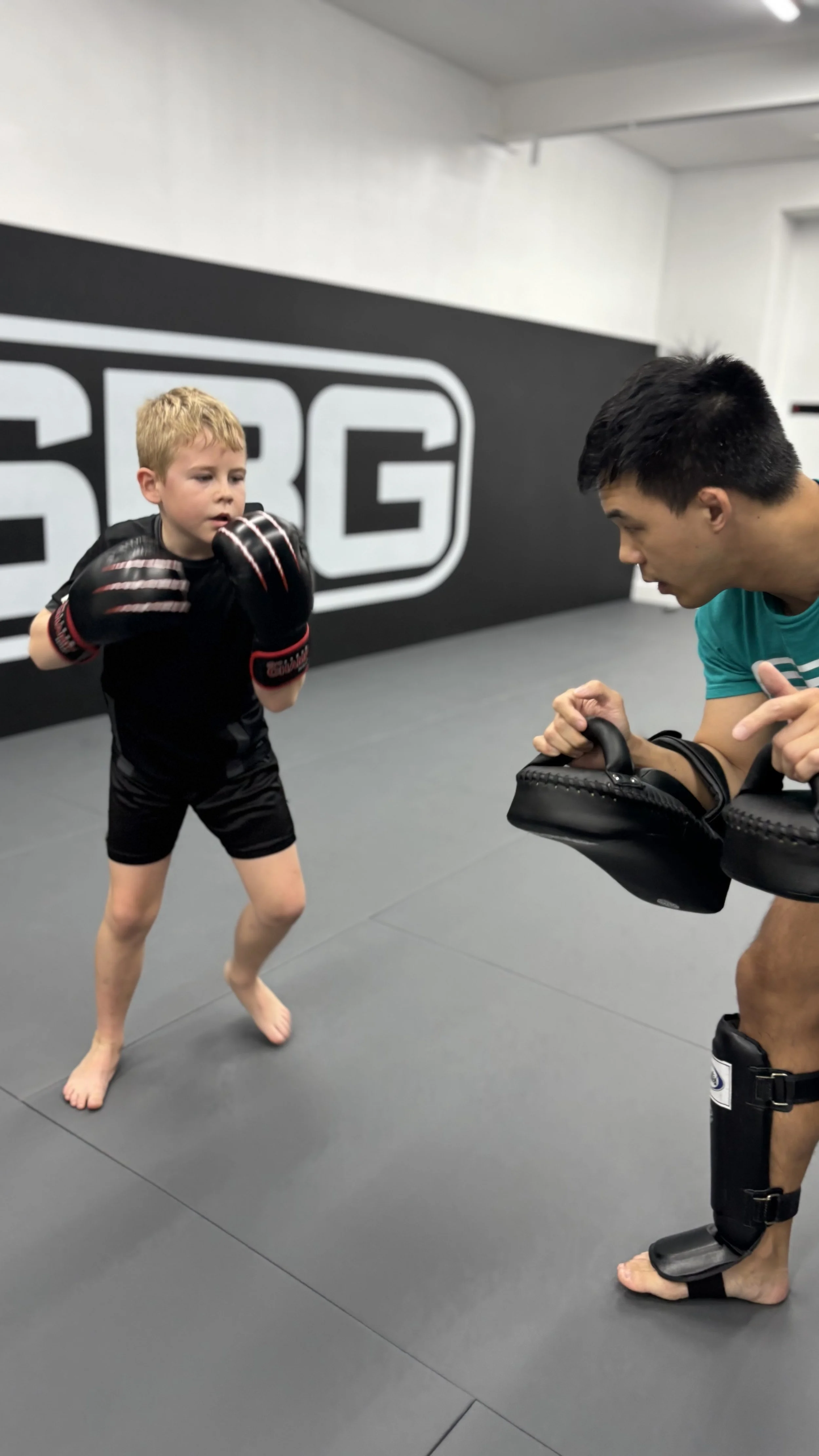 A young boy with blond hair practicing boxing in a gym, facing an adult trainer who is holding focus mitts for him to punch. The boy is wearing black shorts, gloves, and is barefoot. The trainer is wearing a green shirt and black shin guards.