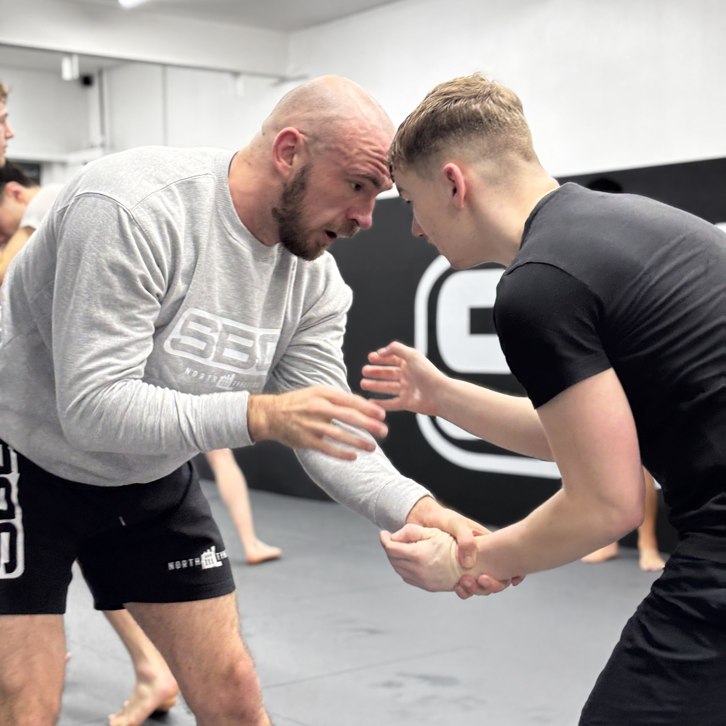Two men practicing Brazilian Jiu-Jitsu in a gym, engaged in a grappling technique, with other practitioners in the background.