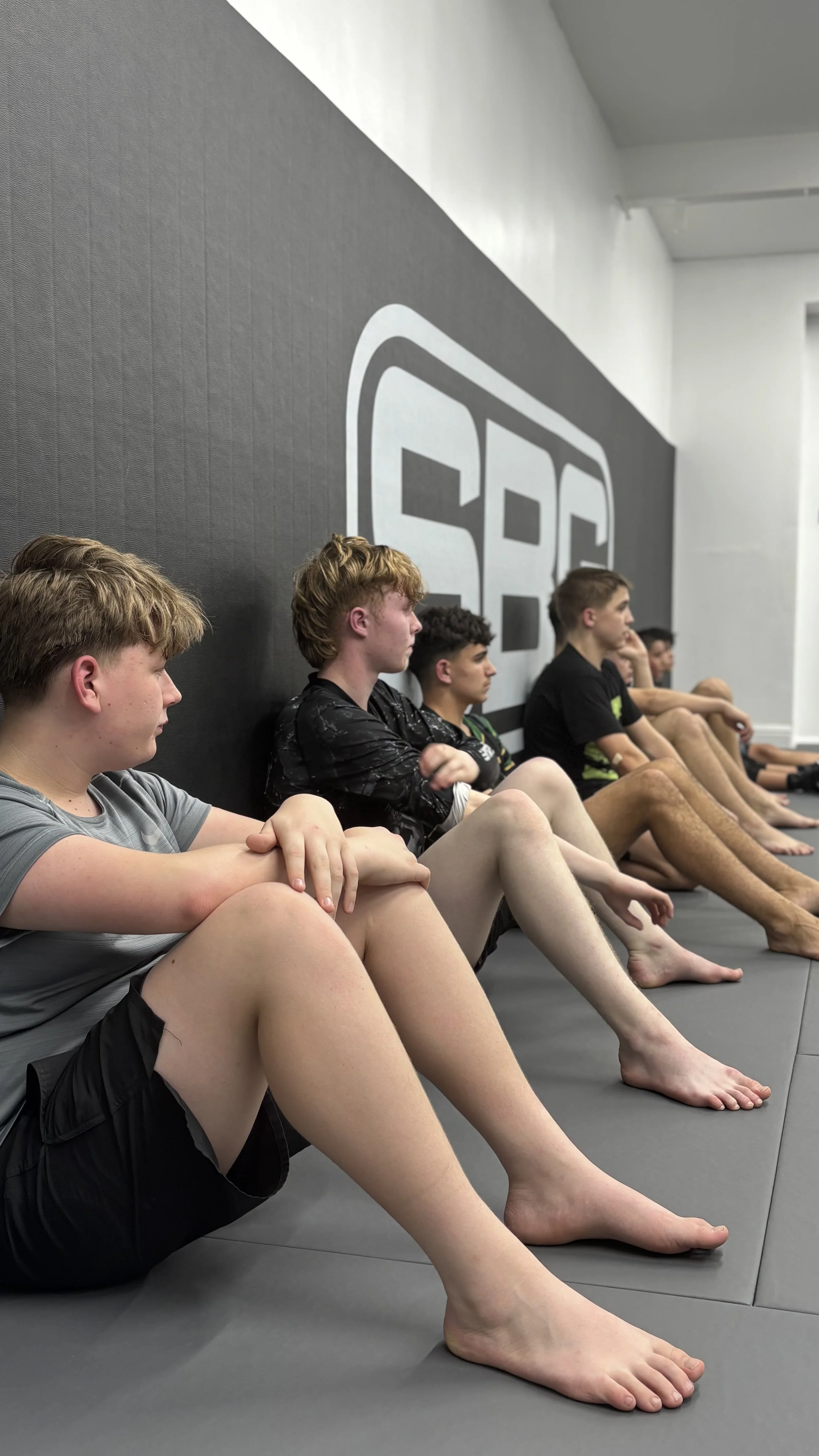 A group of young boys sitting cross-legged on the floor in a martial arts dojo, facing the wall with a large logo that says 'SG'. They are barefoot and appear focused, with some resting their hands on their knees or ankles.