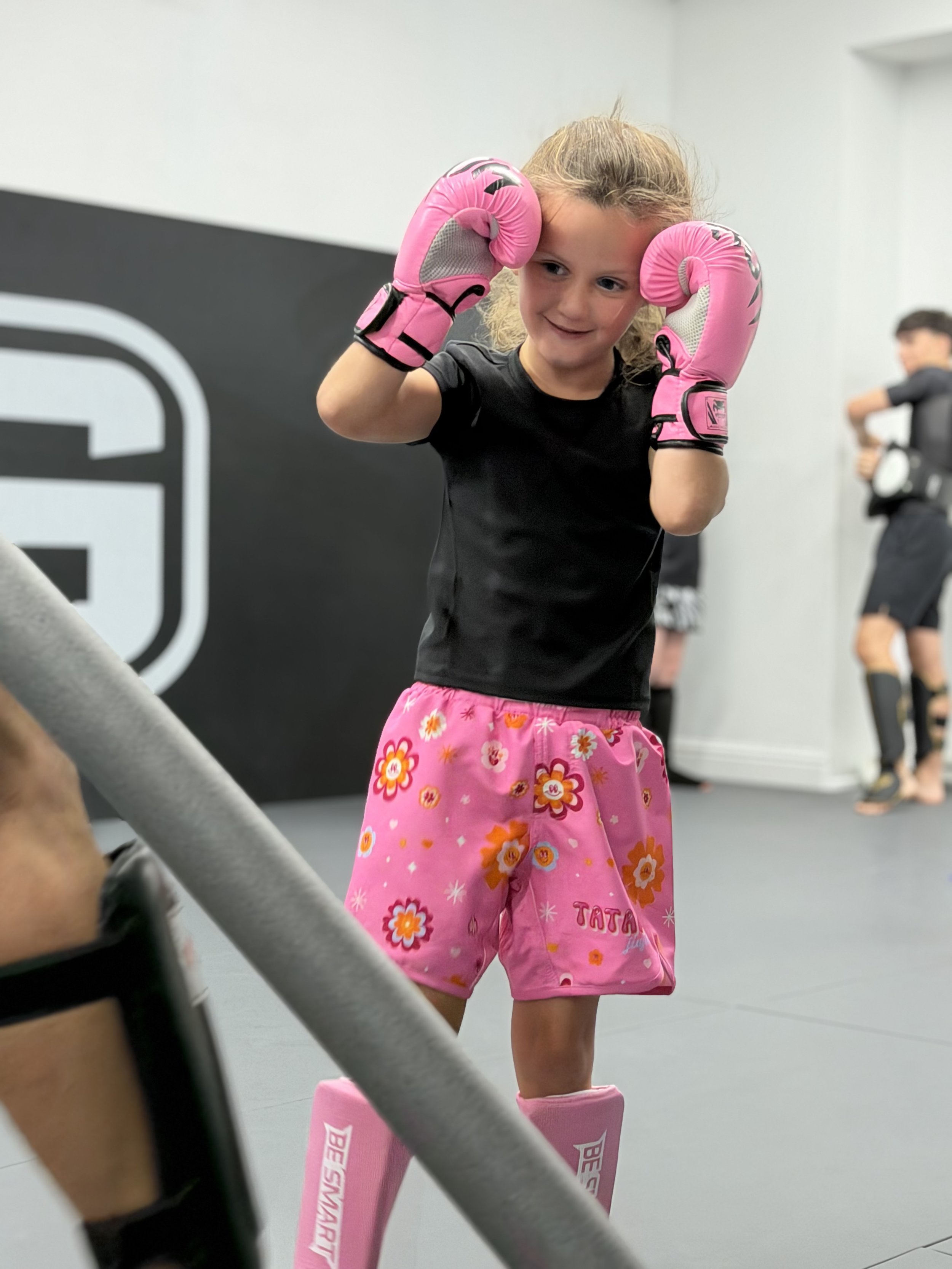 Young girl in black athletic shirt and pink shorts with floral pattern practicing boxing with pink gloves in a gym, smiling while training.