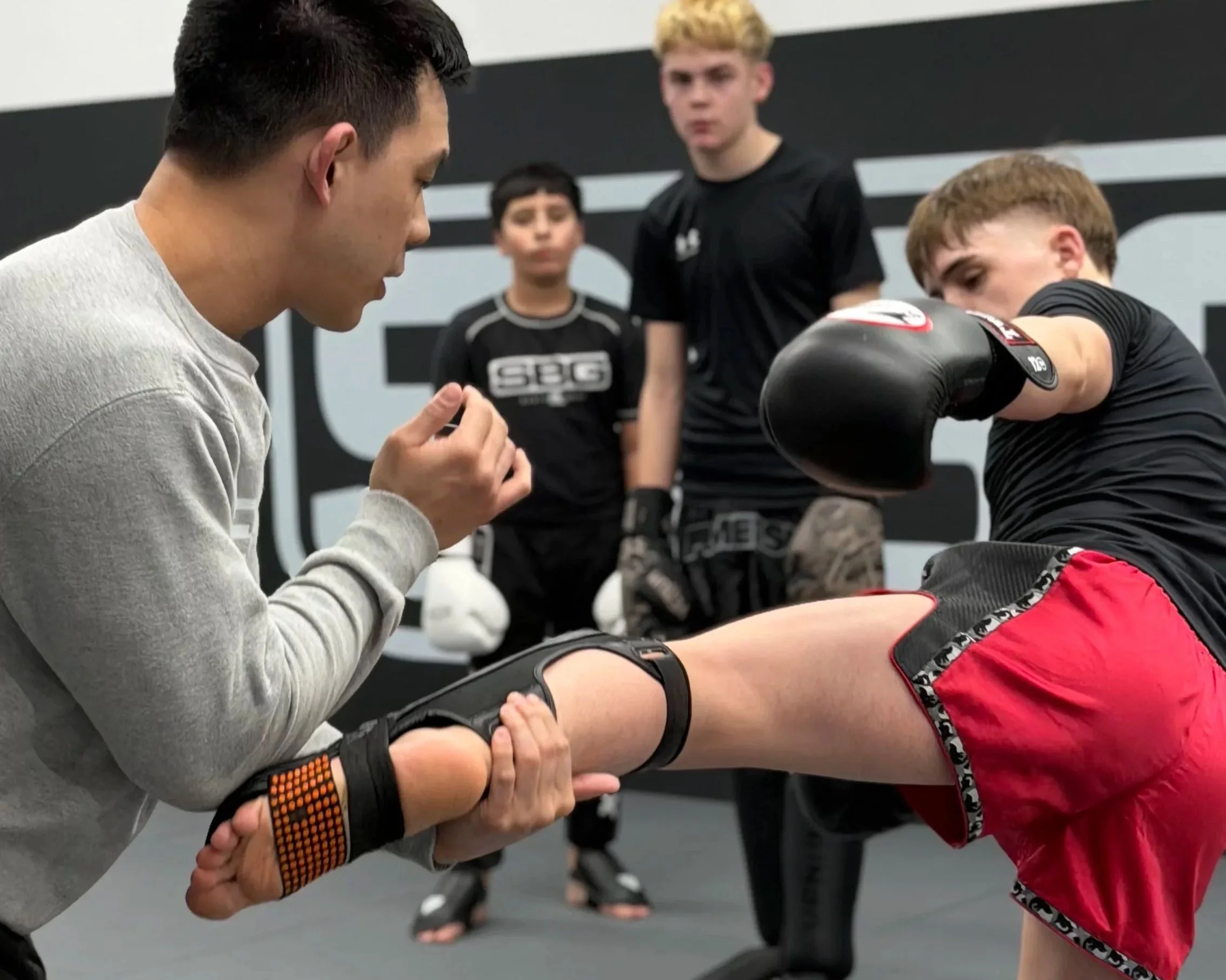 A trainer demonstrates a boxing technique to a young male athlete in a martial arts gym, while three other young trainees observe.