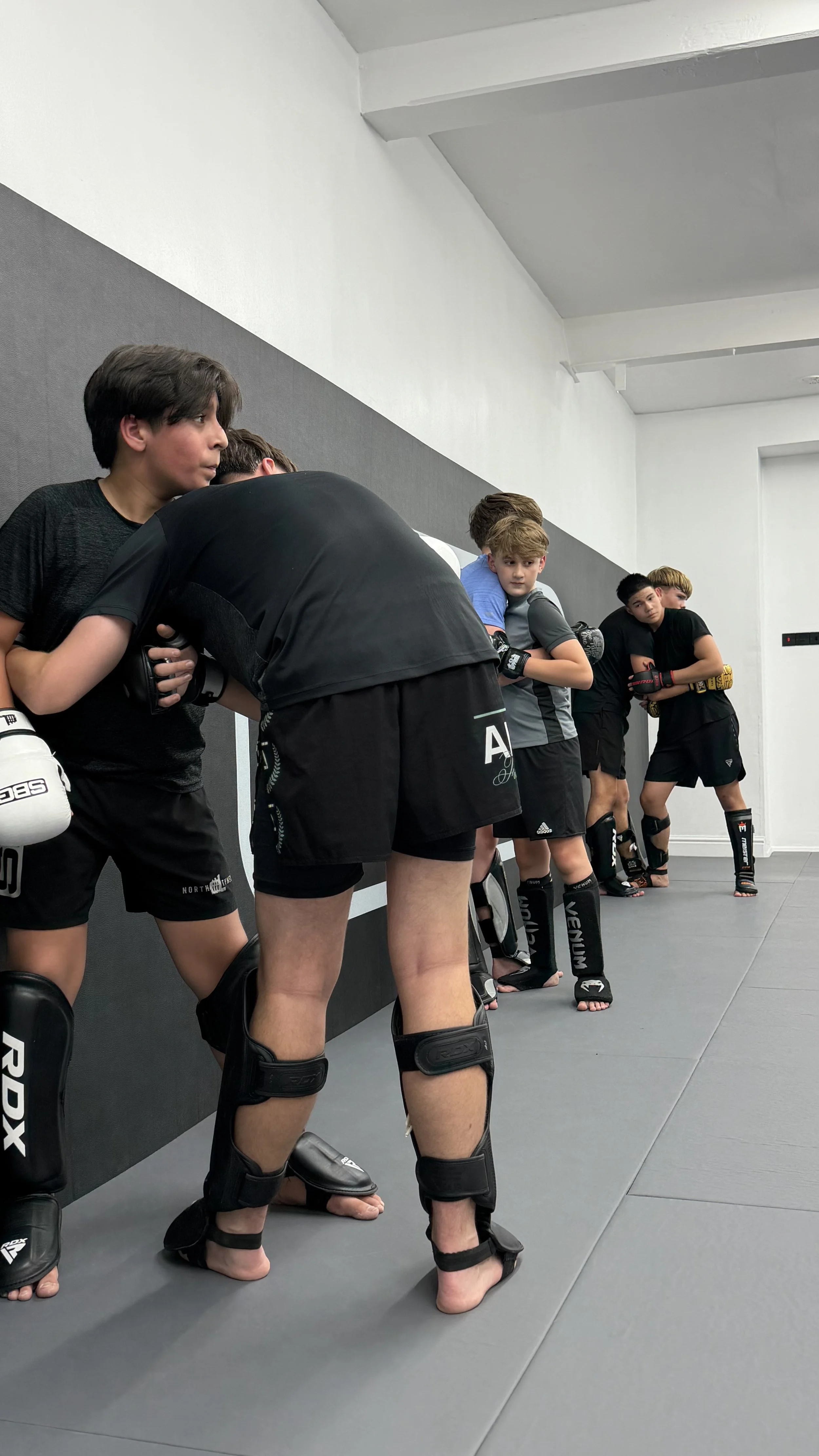 A group of young martial artists or MMA fighters in black gear, including shin guards and gloves, lined up against a wall in a training facility, preparing for a drill or class.