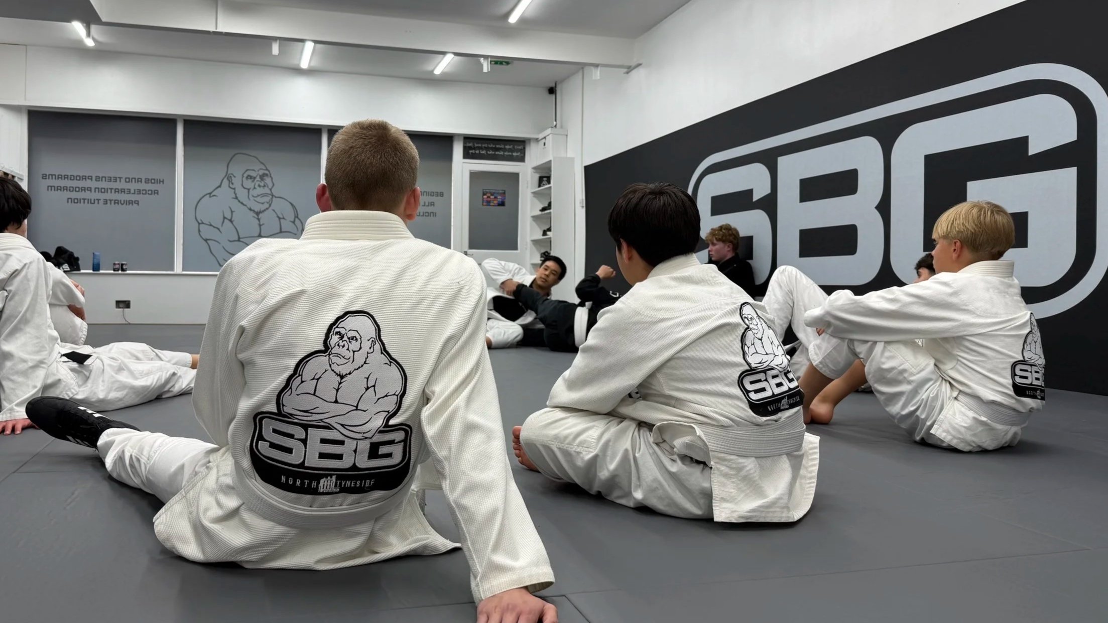 Martial arts students in white uniforms sitting on the floor in a dojo, listening to an instructor who is lying back against the wall, with a large SBG logo on the wall and a cartoon gorilla image.