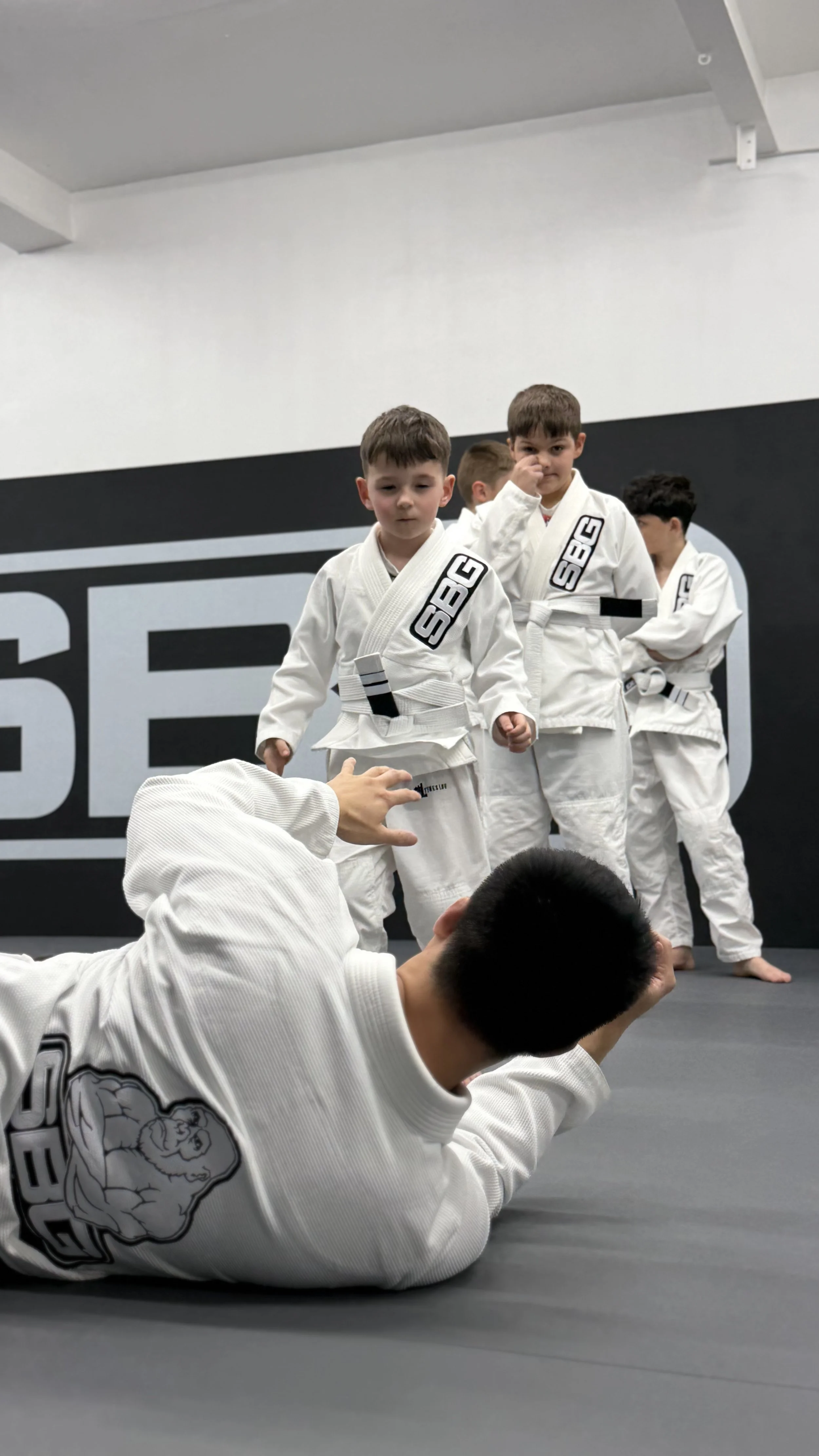 Children in martial arts gi uniforms practicing Brazilian Jiu-Jitsu on a training mat at a dojo.