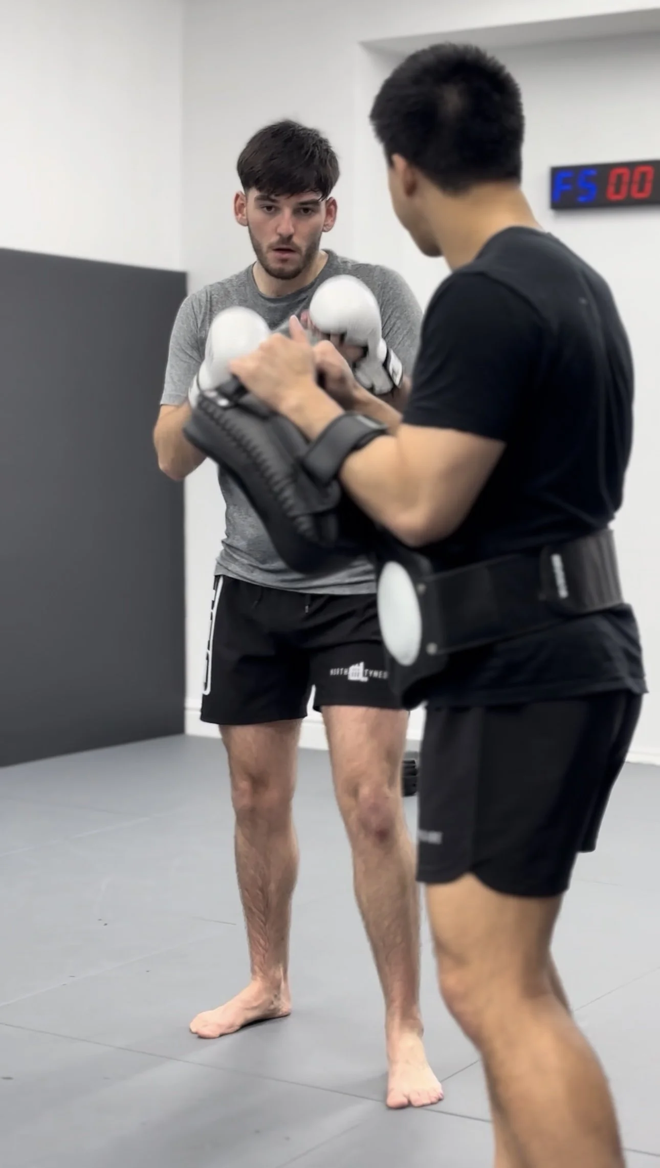 Two men in a martial arts training gym practicing with focus mitts. One man is holding the mitts, and the other is raising his fists in a fighting stance.