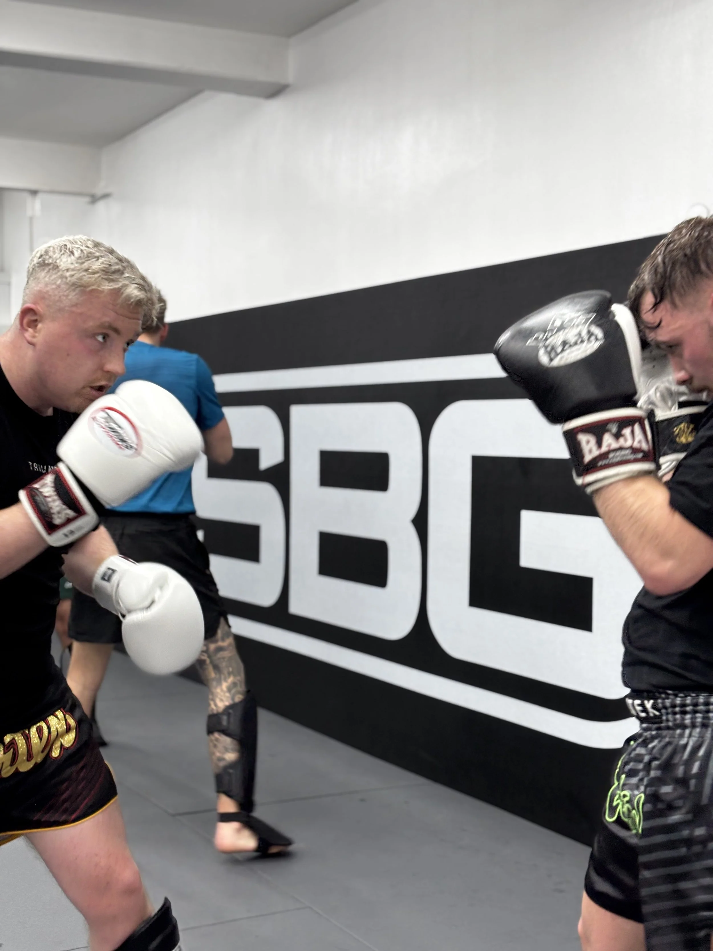 Two men sparring in a boxing gym, facing each other with gloves, in front of a wall with 'SBG' logo.