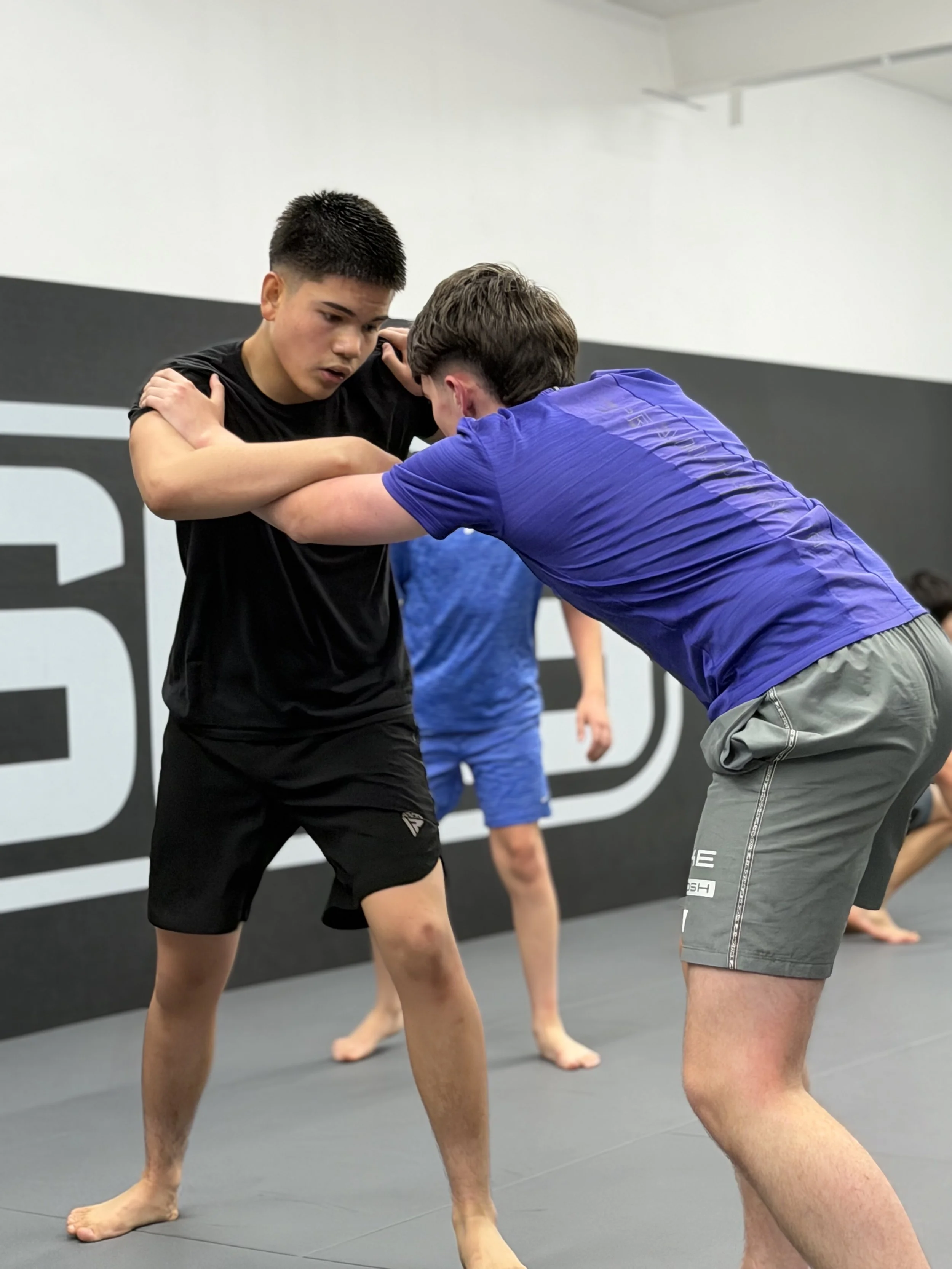 Two men practicing Brazilian Jiu-Jitsu on a gray mat in a gym, with one man in a blue shirt and gray shorts and the other in a black shirt and black shorts, demonstrating a grappling technique.