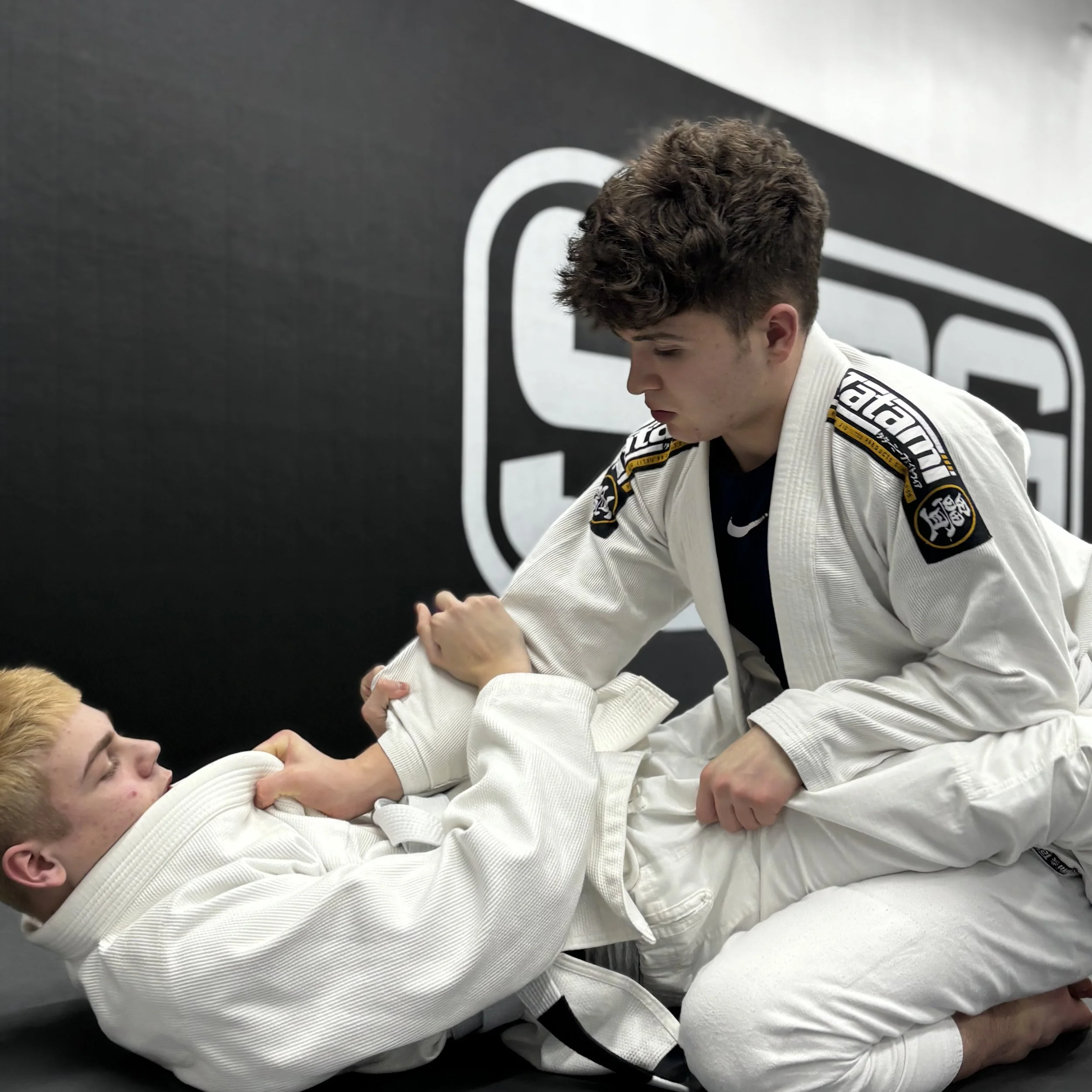 Two young men in white jiu-jitsu gis with black belts practicing grappling on the mat, with one man gripping the other's sleeve while the other is positioned in a dominant stance.