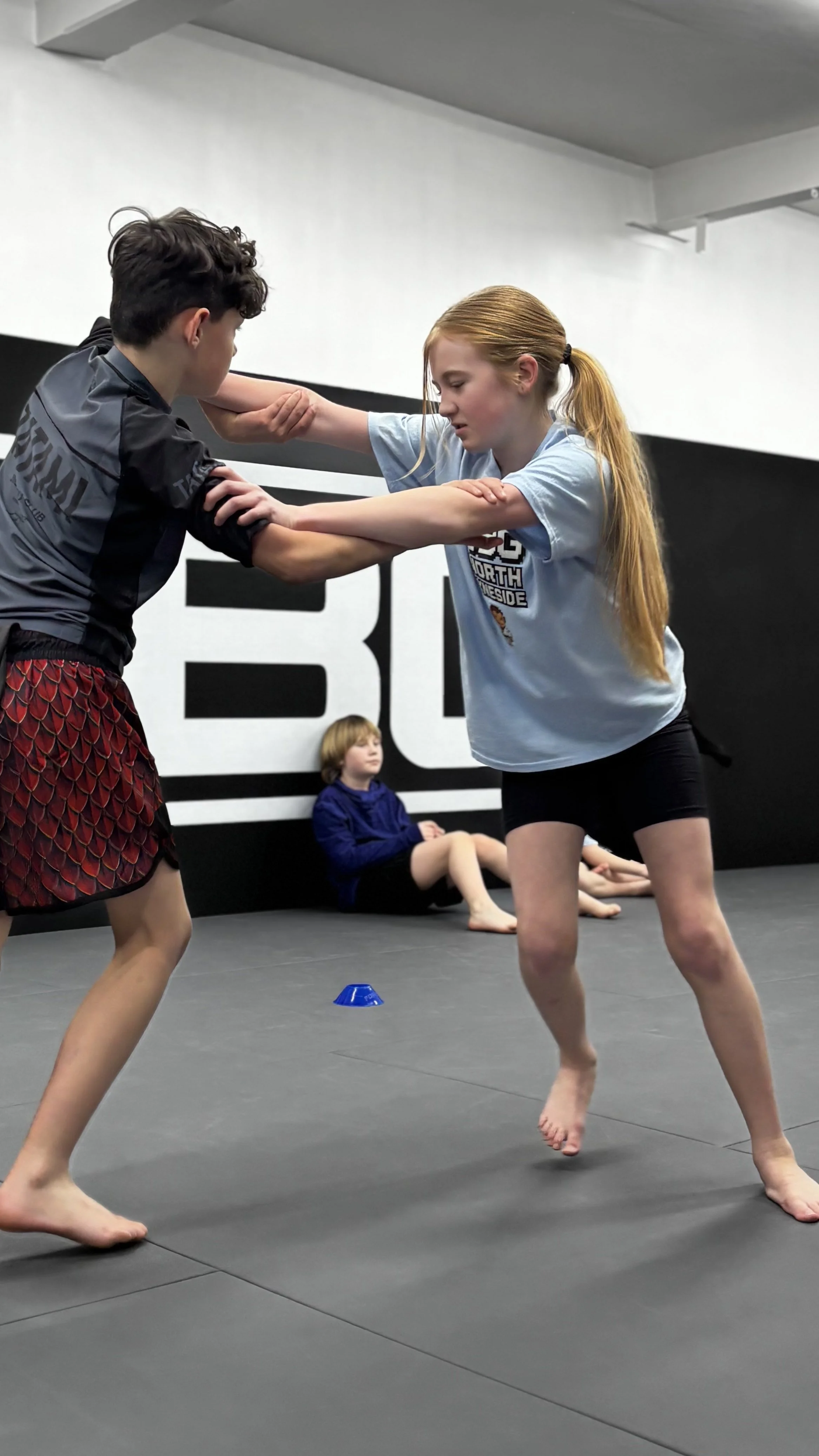 Two children practicing Wrestling on a black mat in a gym, with a third kid sitting in the background.