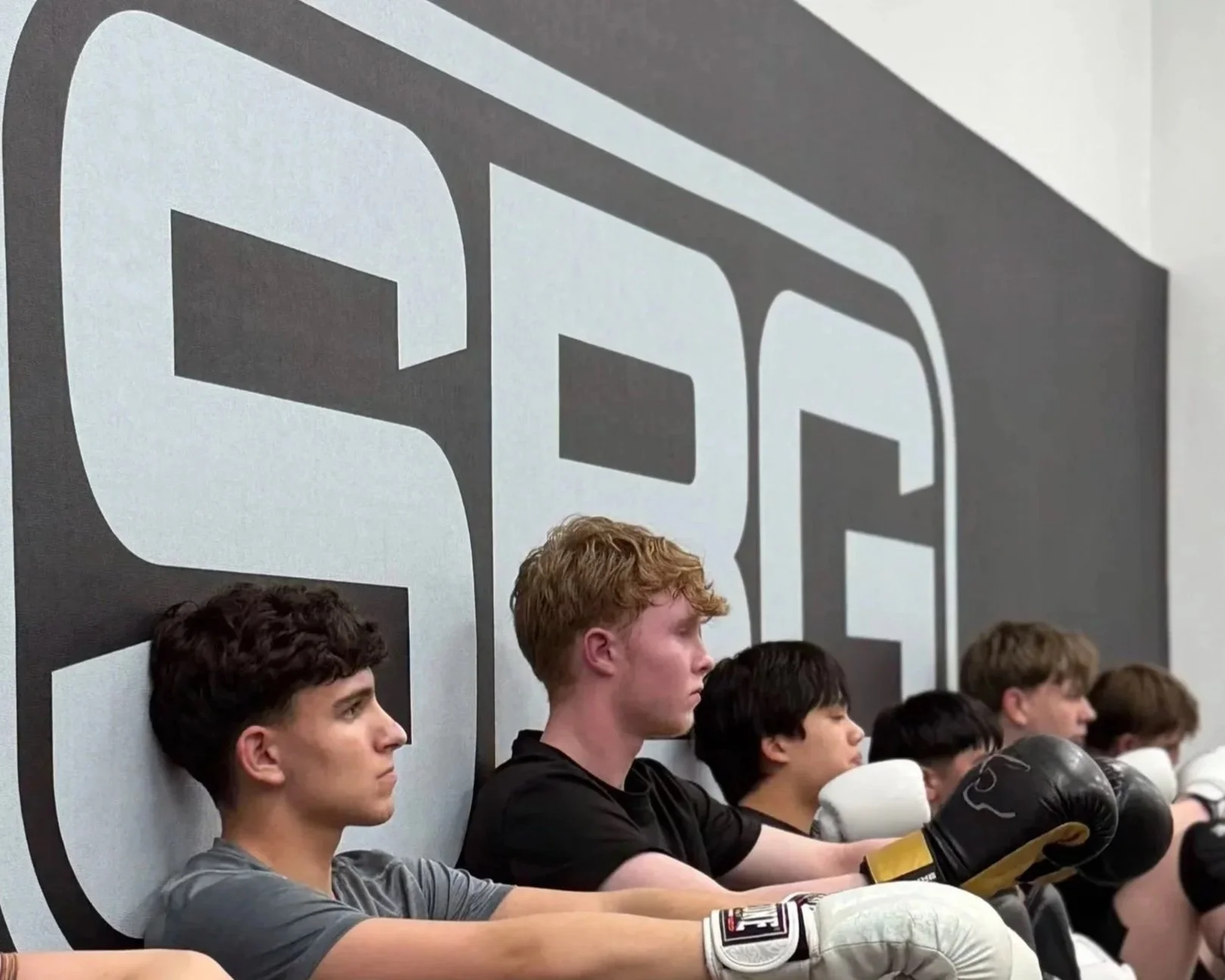 A group of young men sitting against a wall with a large black and white fight logo behind them, some wearing boxing gloves.