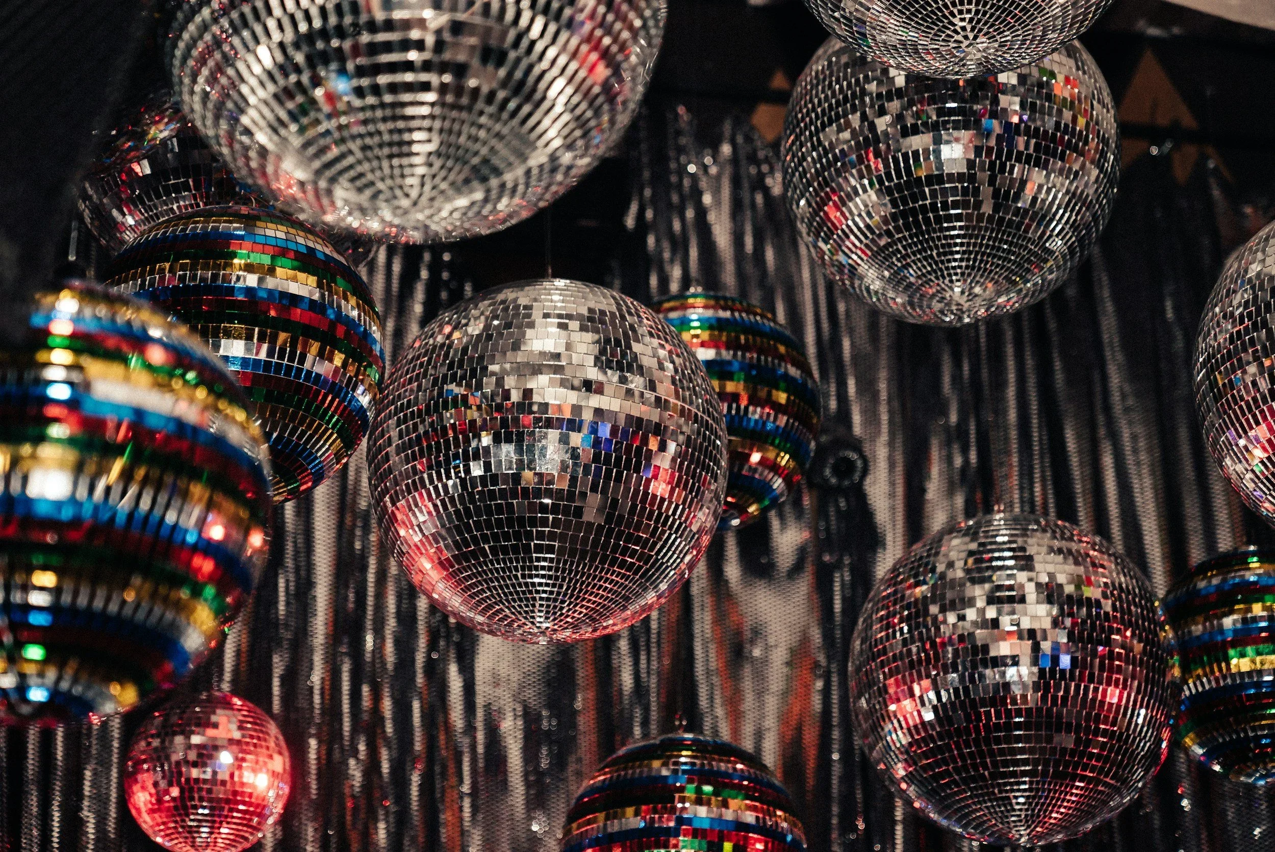 Multiple disco balls hanging from a ceiling with reflective, mirrored surfaces and a black backdrop.