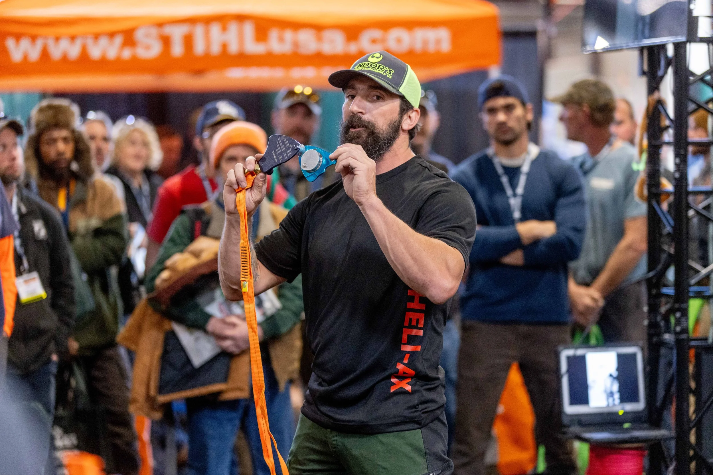 A man with a beard wearing a black shirt and a green cap with yellow accents, holding a rigging device, speaking to a crowd at an outdoor event with a blurred background of people and a banner.