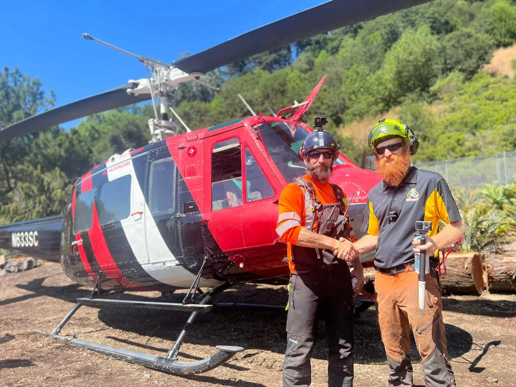 Two rescue workers shake hands in front of a red disaster response helicopter with black blades, surrounded by green trees and a clear blue sky.