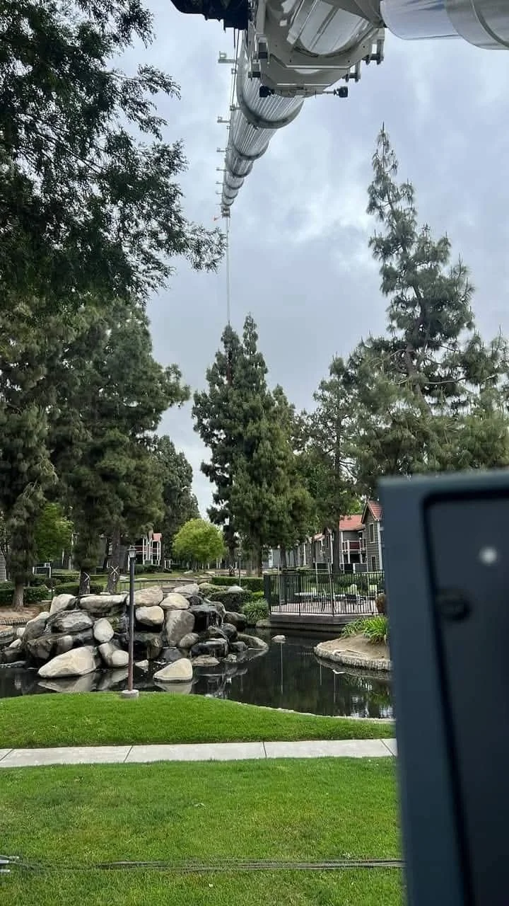 Photo taken from below a tall crane or lift, looking up at its arm extending into a cloudy sky over a park with large trees, rocks, a pond, and houses in the background.