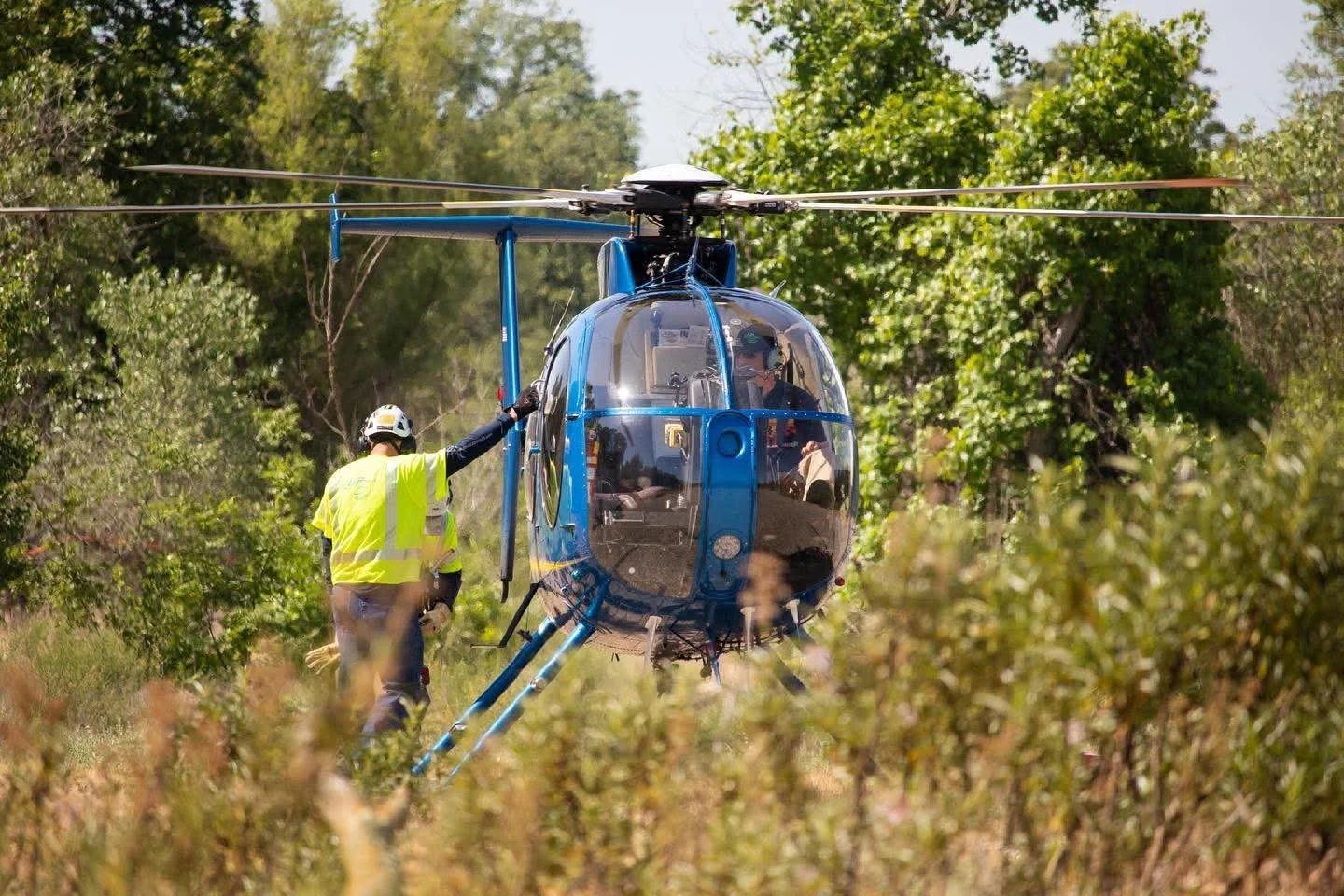 A blue helicopter on a grassy field surrounded by trees, with a person in a yellow safety vest and helmet standing next to it, holding onto the helicopter.