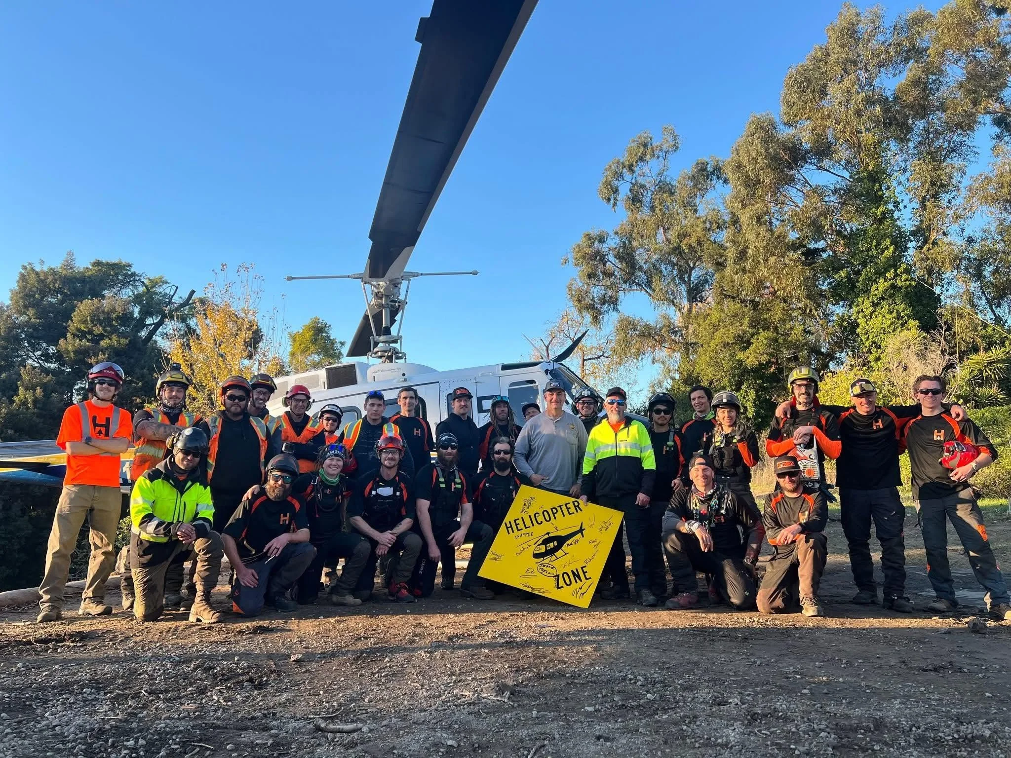 Group of people outdoors at a helipad, posing in front of a helicopter, with trees and a bright blue sky in the background. Some are wearing helmets and safety gear.