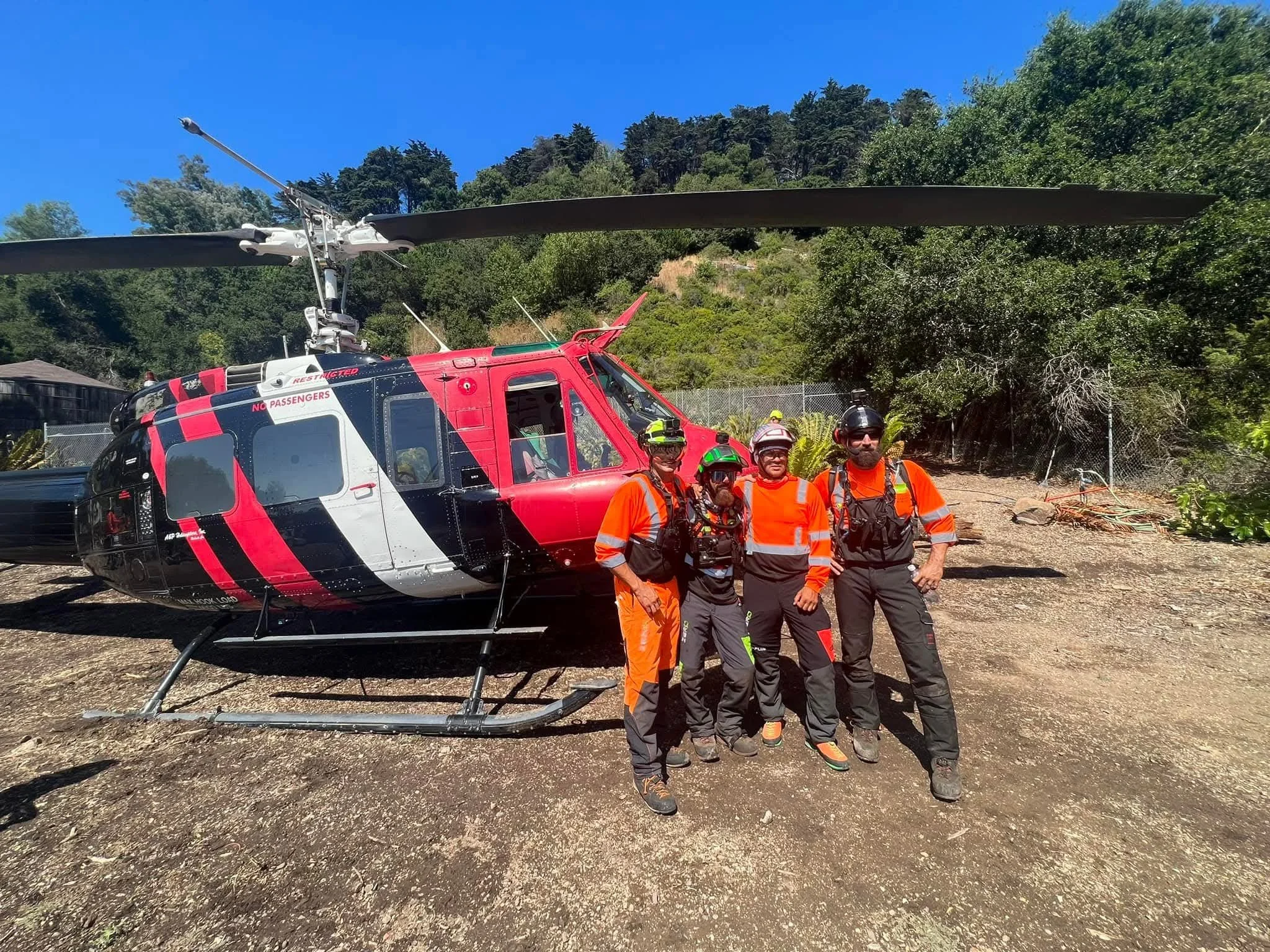 Four rescue team members wearing helmets and orange safety vests standing in front of a red and white rescue helicopter on a dirt area with trees and a fence in the background.