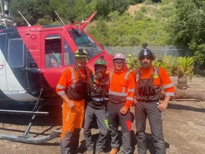 Four tree workers in orange uniforms and hats standing next to a red helicopter outdoors with greenery in the background.