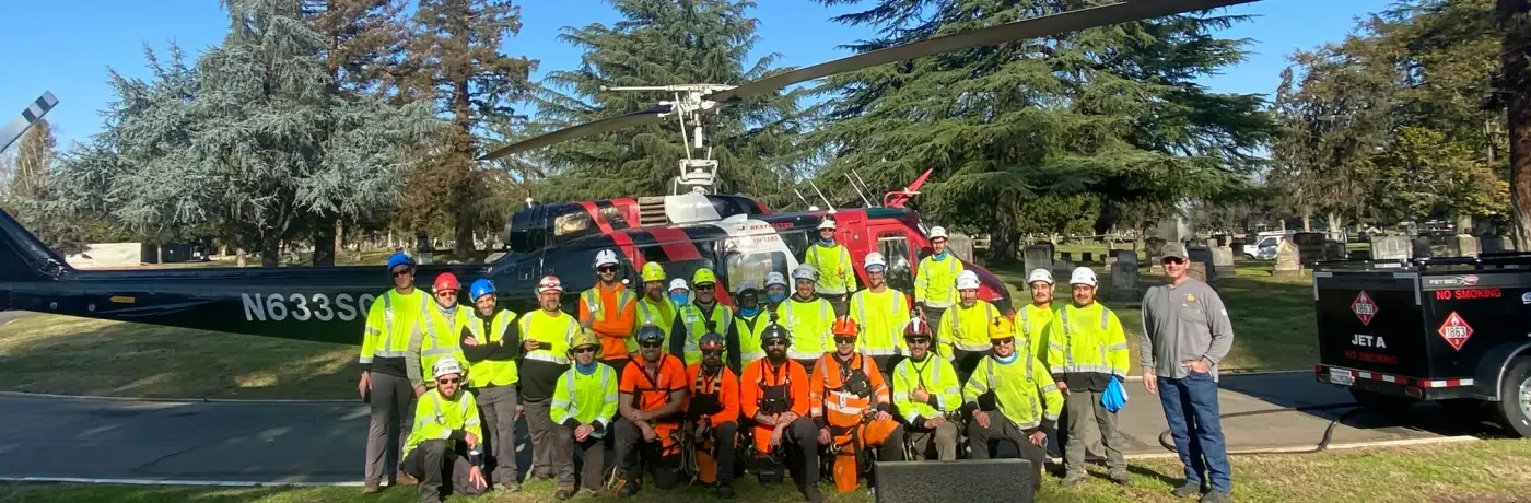 Group of helicopter  team members posing in front of a helicopter and  equipment in a park with trees.