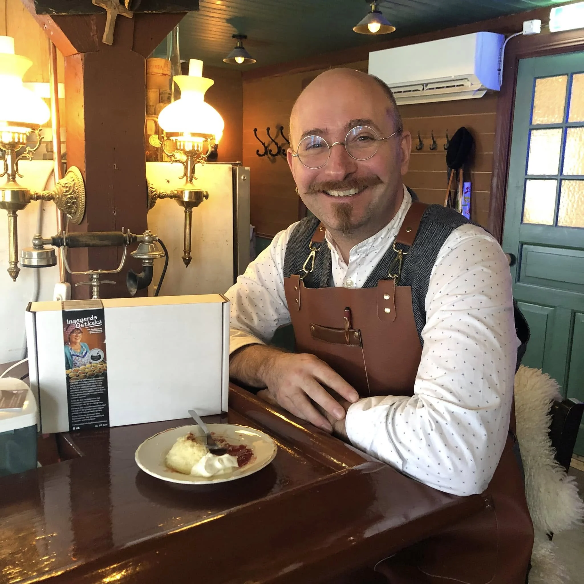 Glad man med skägg och mustasch sitter vid ett bord i en rustik inredning, med fikabord, bakgrundsbelysning och en tallrik med mat.