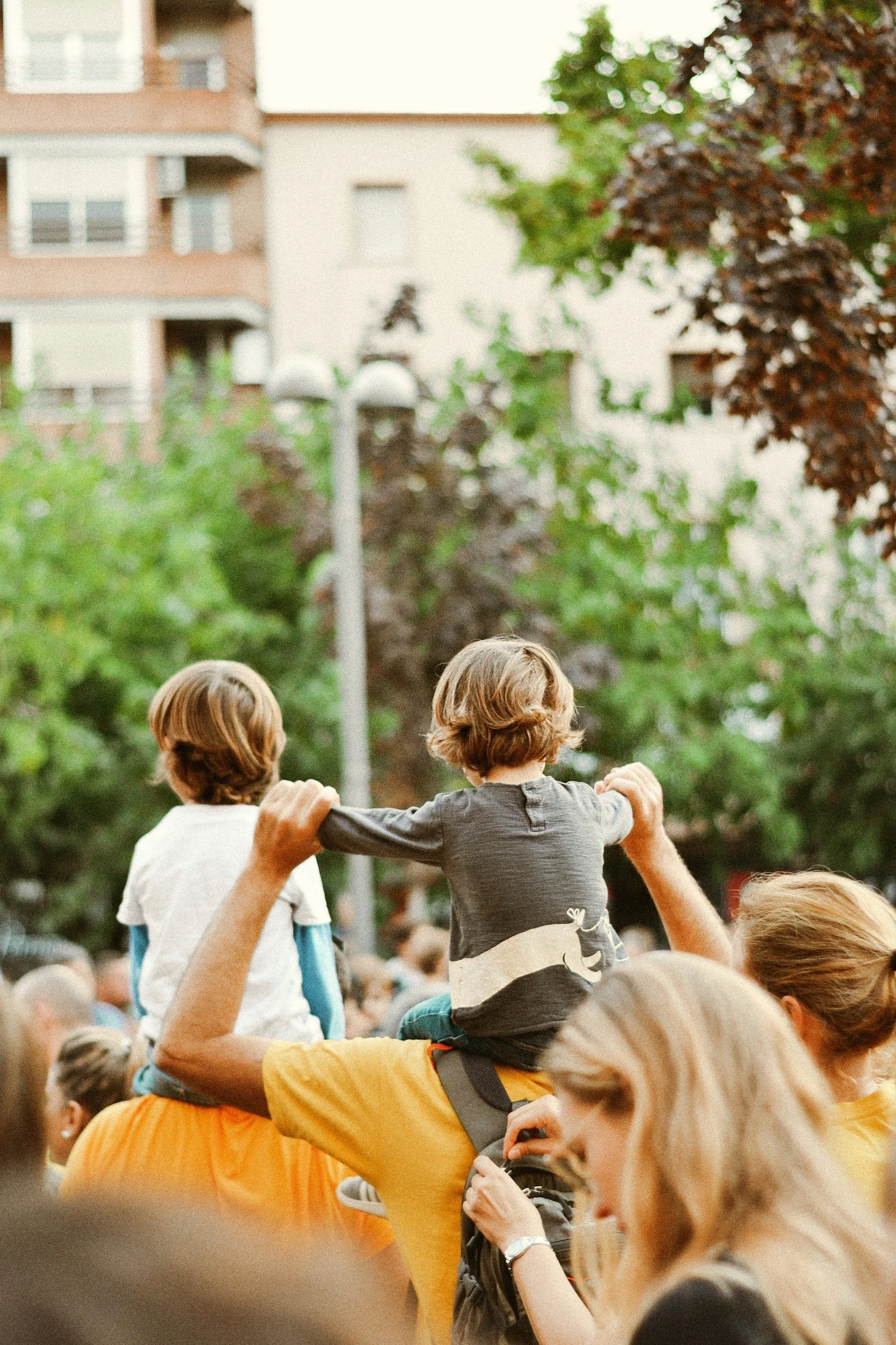 Children sitting on shoulders at an outdoor event during daytime, surrounded by adults and other children, with trees and buildings in the background.